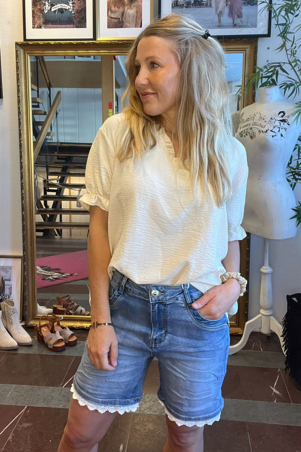 Kvinna i vit blus och jeansshorts står med ena handen i fickan och tittar åt sidan. Hon poserar i en butikslik miljö med stor guldspegel, skor på golvet och provdocka med texten ”Fanny Michel”.