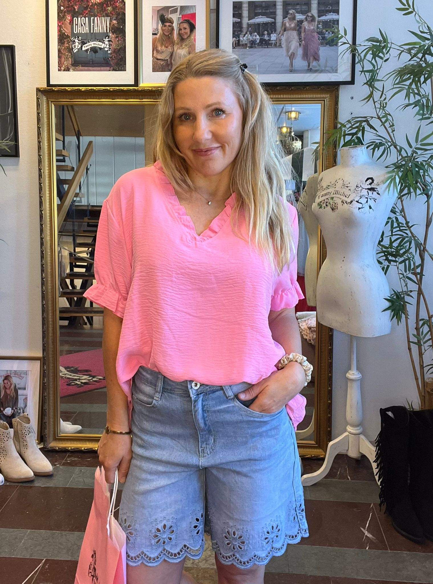 Leende kvinna i rosa blus och blå shorts står med handen i fickan och håller en rosa påse. Bakom syns spegel, inramade foton med texten “CASA FANNY” och en provdocka med “Fanny Michel”.