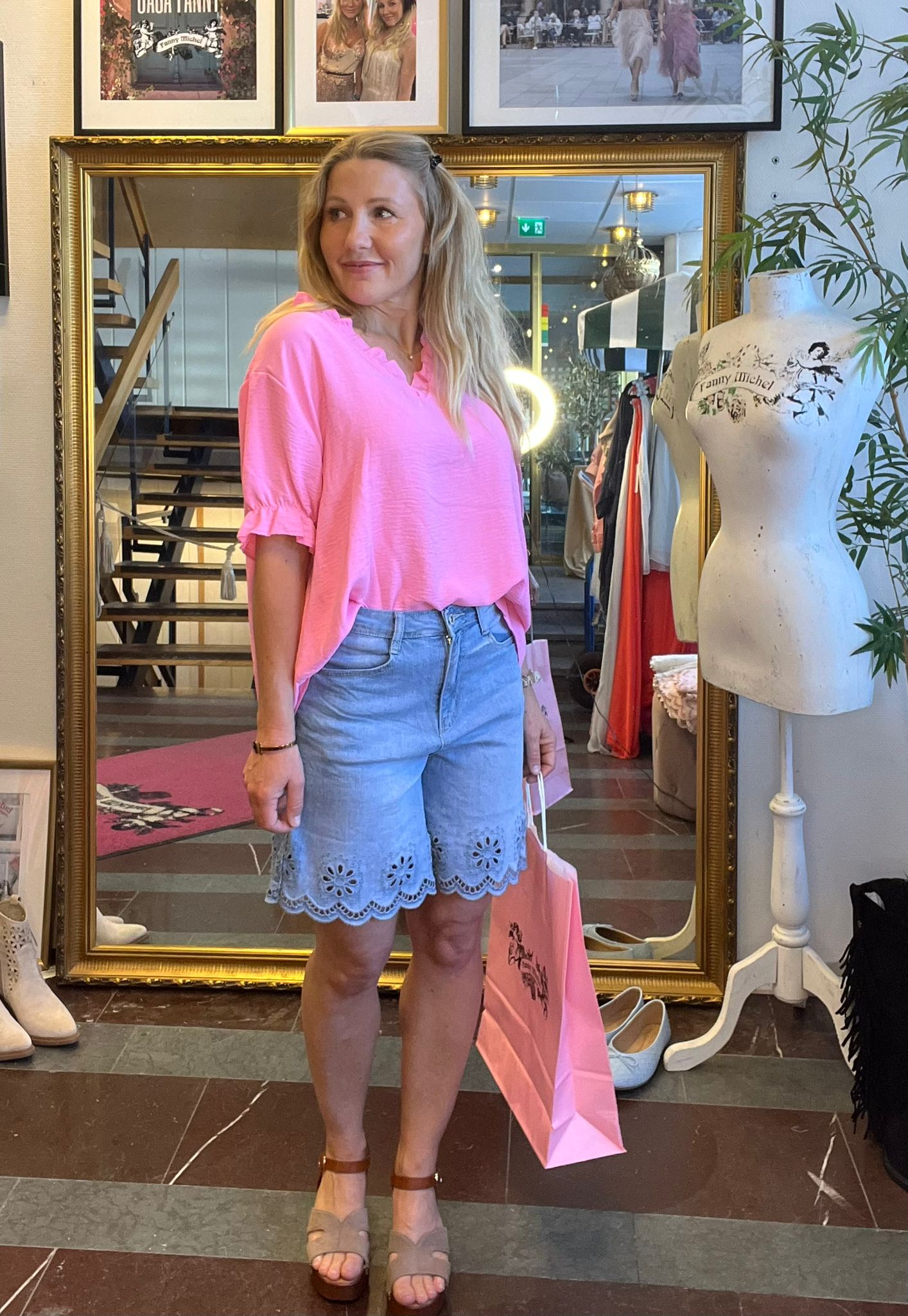 Kvinna i neonrosa blus och ljusblå spetskantade shorts står och tittar åt sidan i en klädbutik, håller rosa shoppingpåse. Bakom henne syns stor guldspegel, klädställning och provdocka med texten “Tanny Michel”.