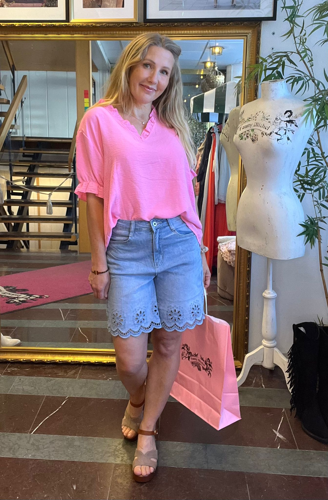 Leende kvinna står i en klädbutik och håller en rosa shoppingpåse, klädd i rosa topp och ljusblå shorts med spetskant. Bredvid syns en provdocka med texten “Fanny Michel” och klädställningar i bakgrunden.
