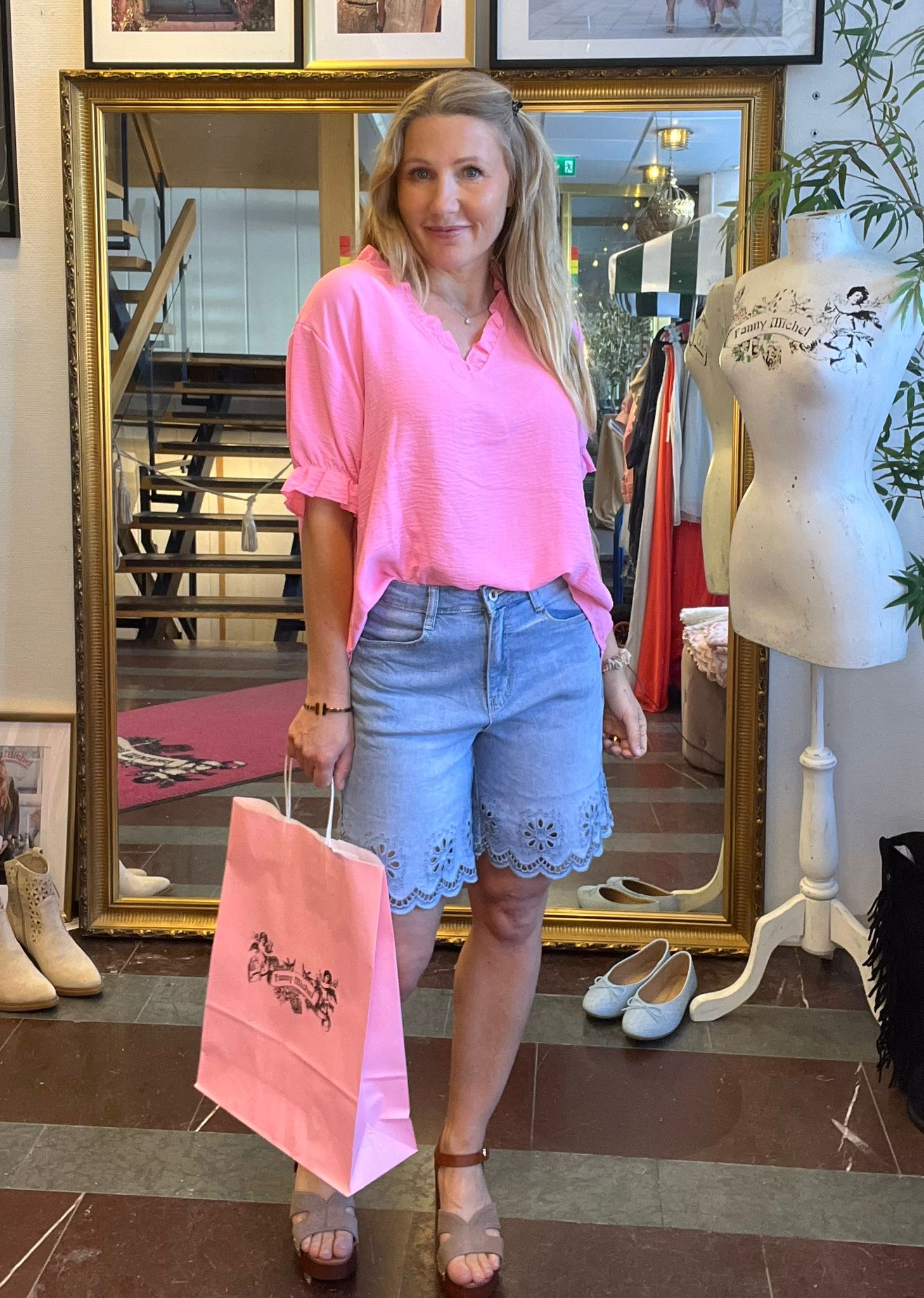 Kvinna i rosa blus och ljusblå shorts står och ler framför en stor guldramsspegel, med en rosa shoppingpåse i handen. Runtom syns kläder, skor och en provdocka med texten “Family Affair”.