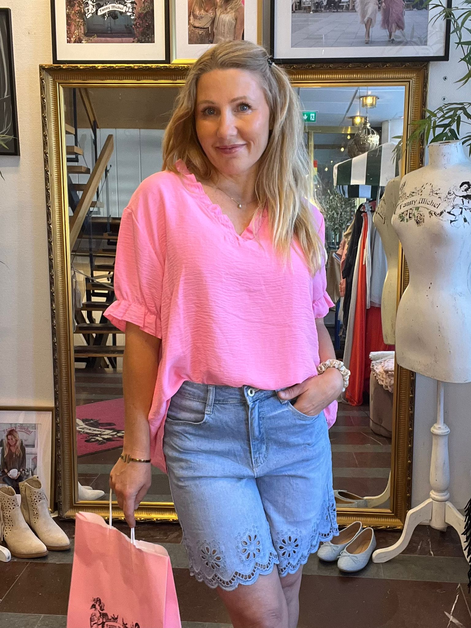 Blond kvinna i rosa blus och ljusblå shorts står och ler i en klädbutik, med ena handen i fickan och en rosa shoppingpåse i den andra. Bakom syns stor spegel, klädställning och skyltdocka med texten ”Fanny Michel”.