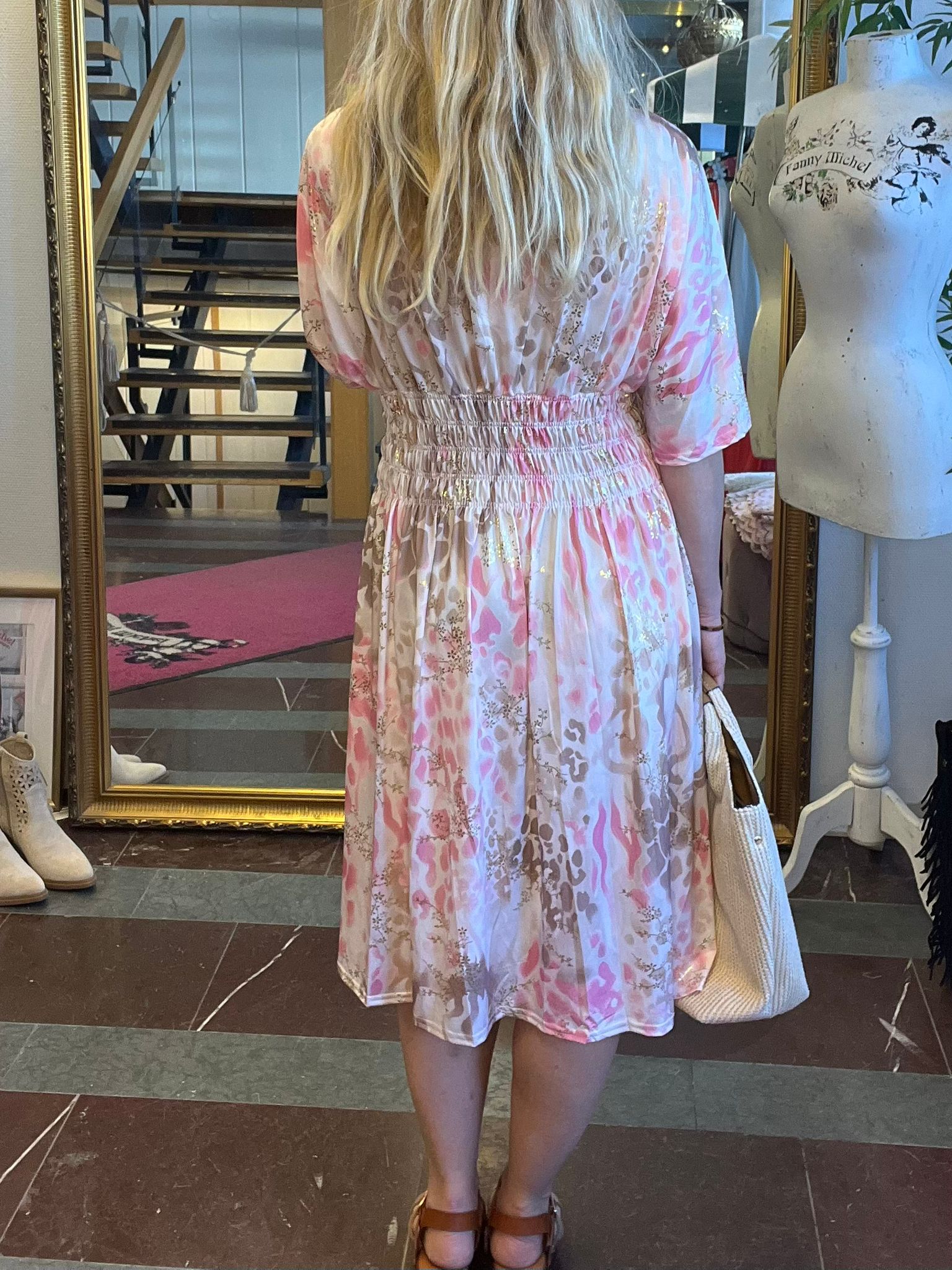 Person med långt blont hår står med ryggen mot kameran och håller en vit tygväska, iklädd rosa- och beige mönstrad klänning med smock i midjan. Inomhus vid stor guldramsspegel, trappa och provdocka med texten ”Fanny Michel”.