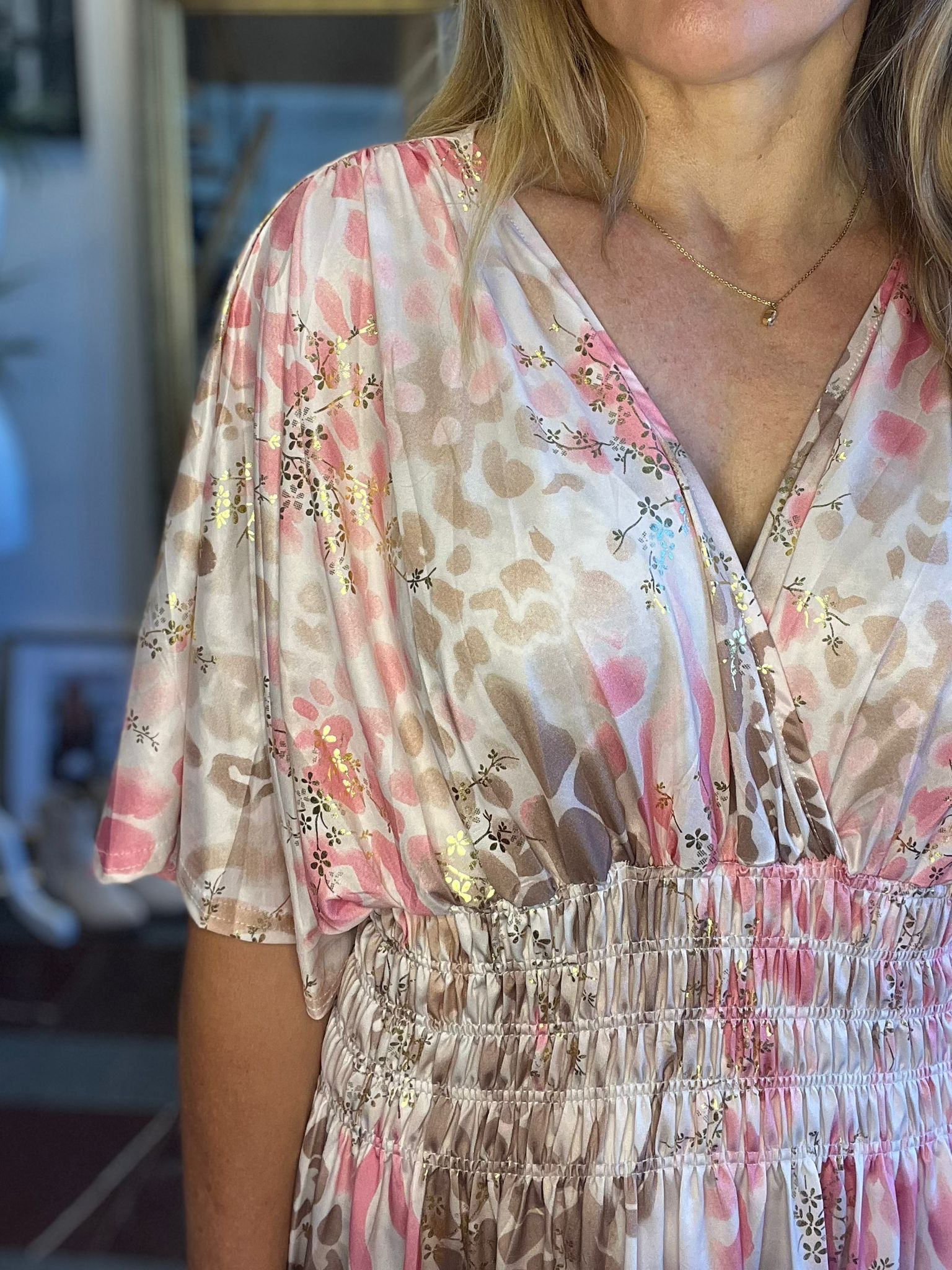 Kvinna står inomhus och visar överdelen av en ljus, V-ringad klänning med puffärmar, rosa och beige mönster samt gulddetaljer. Hon bär ett tunt halsband med liten berlock; bakgrunden är suddig.