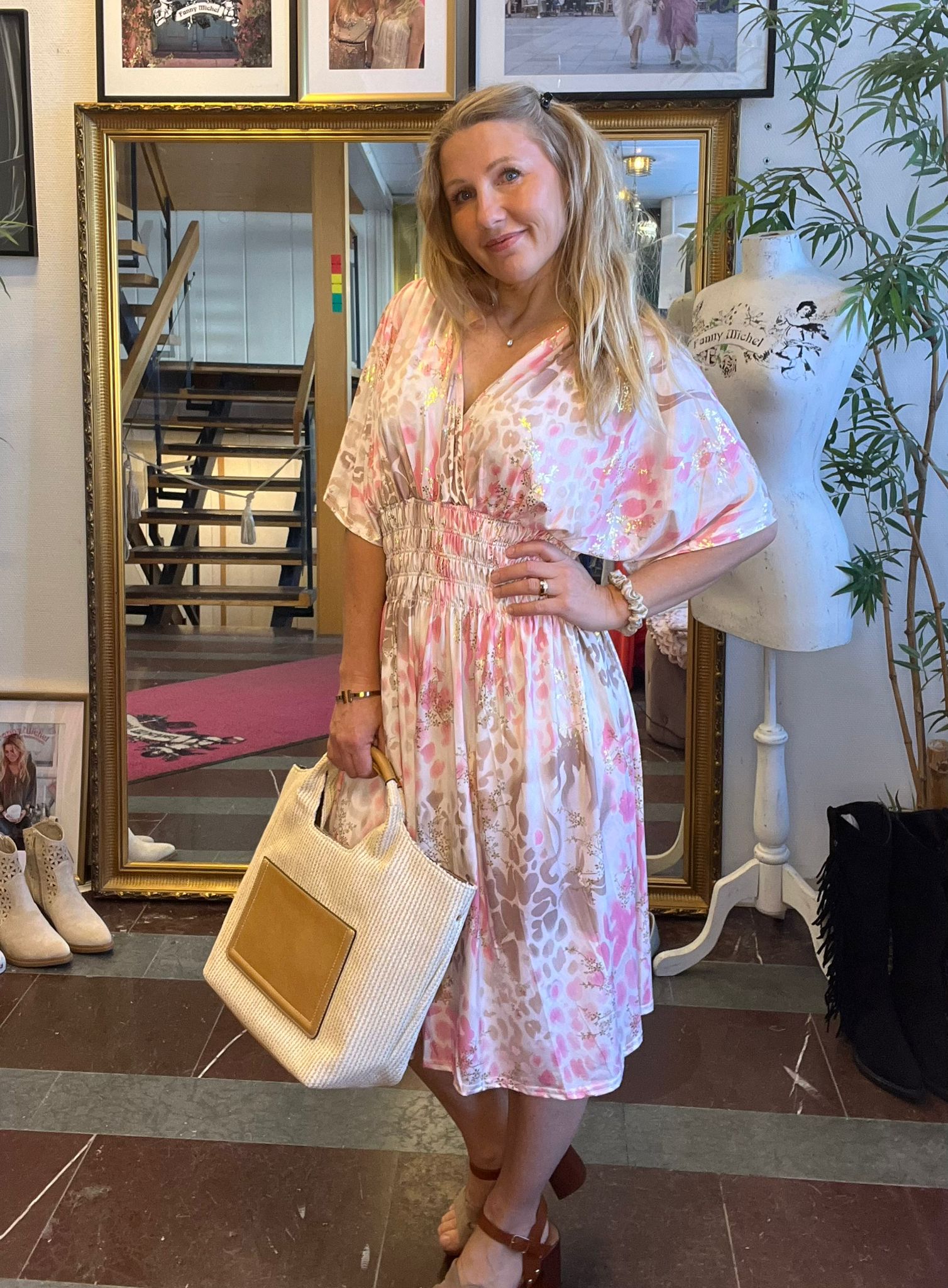 Leende kvinna poserar med handen på höften och håller en stor stråväska, klädd i rosa mönstrad klänning. Hon står i en butik framför en guldramsspegel, bredvid en provdocka med texten “Fanny Michel”.