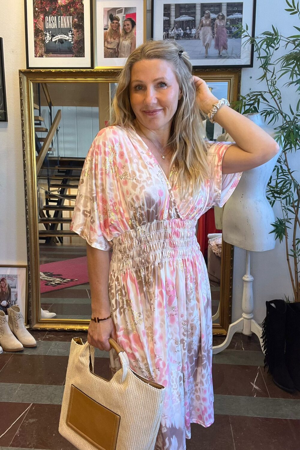 Leende kvinna i rosa och vit mönstrad klänning poserar med ena handen i håret och håller en stor tygväska. Hon står framför en guldramad spegel i en butik med skor, skyltdocka och växt; text: “CASA FANNY”.