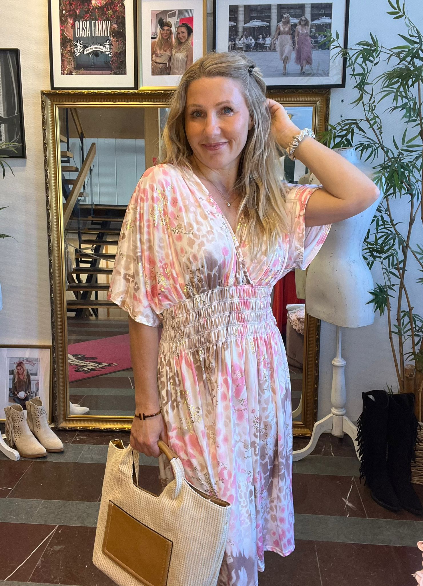 Leende kvinna i rosa och vit mönstrad klänning poserar med ena handen i håret och håller en stor tygväska. Hon står framför en guldramad spegel i en butik med skor, skyltdocka och växt; text: “CASA FANNY”.