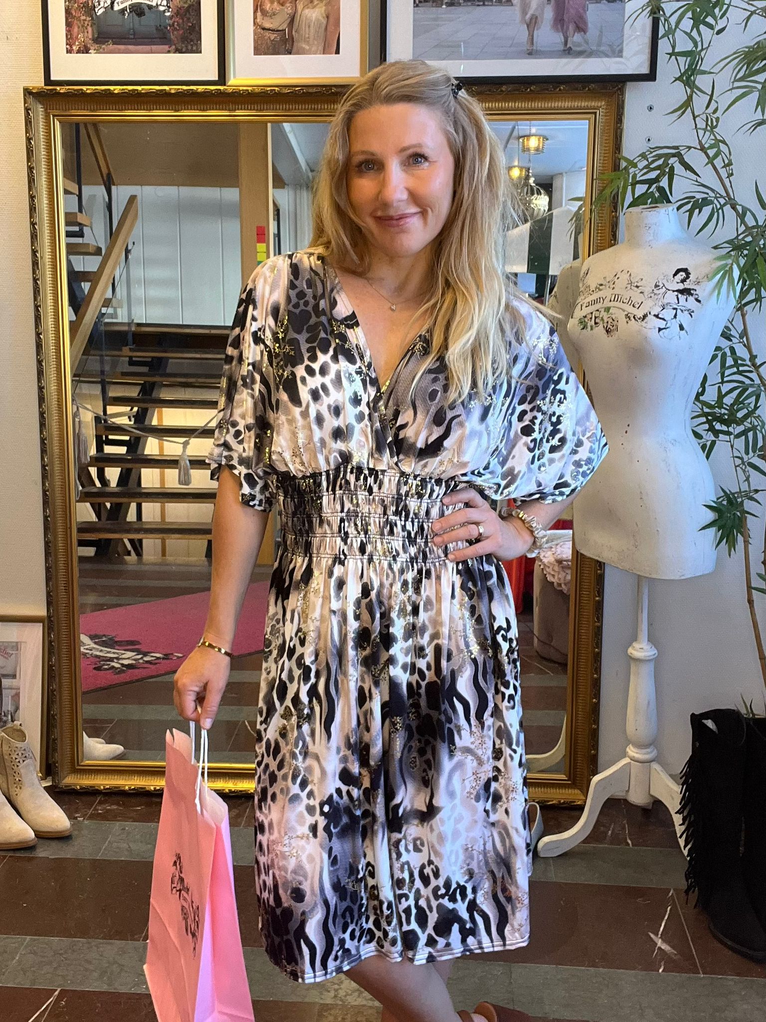 Leende kvinna står med handen på höften och håller en rosa shoppingpåse, klädd i mönstrad klänning. Hon står framför en stor guldramad spegel i en butik; bredvid syns en provdocka med texten “Funny Michel”.
