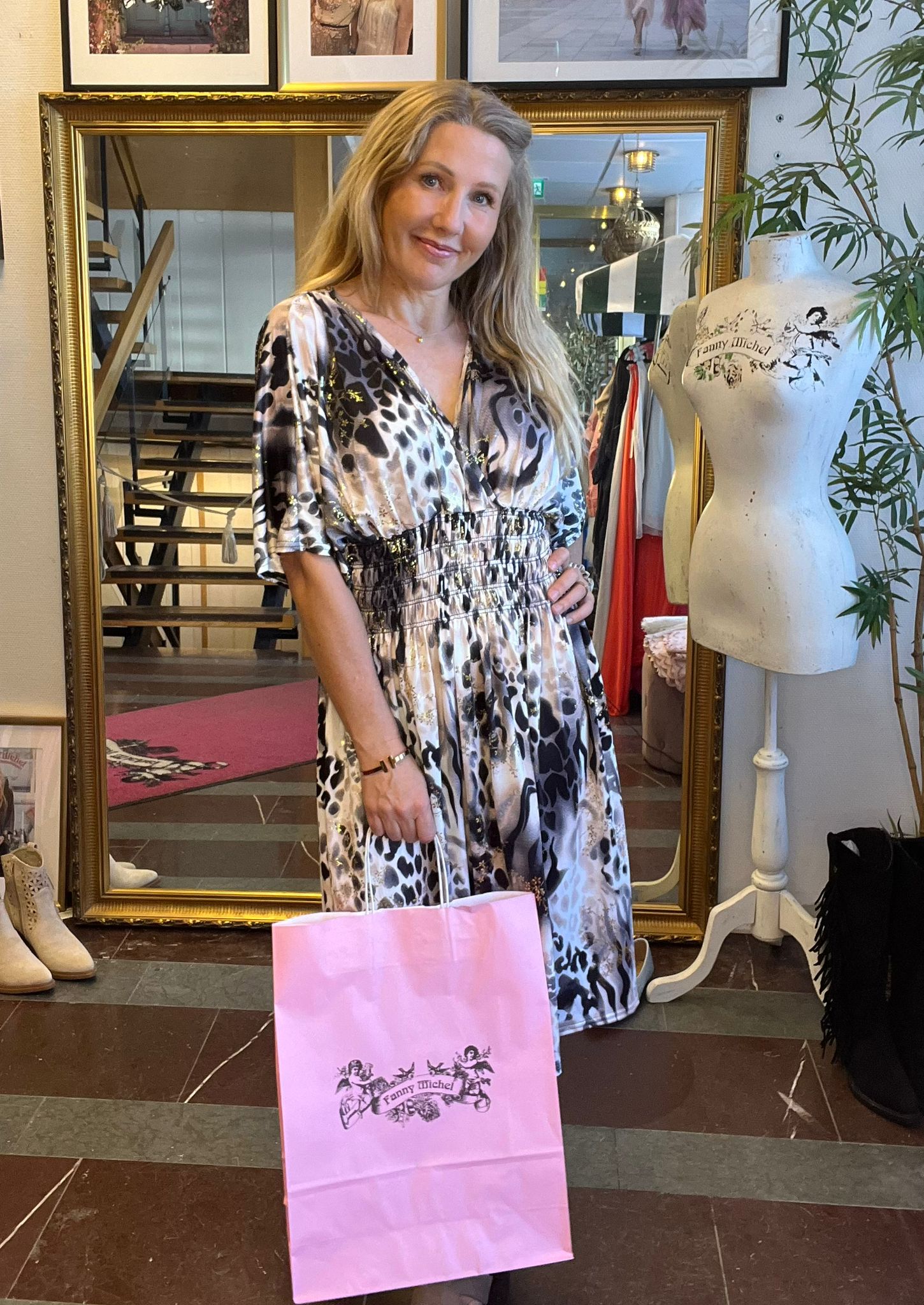Blond kvinna står och ler i en butik, håller en rosa shoppingpåse med texten ”Fanny Michel”. Hon bär mönstrad klänning och poserar framför en stor guldramsspegel bredvid en provdocka.