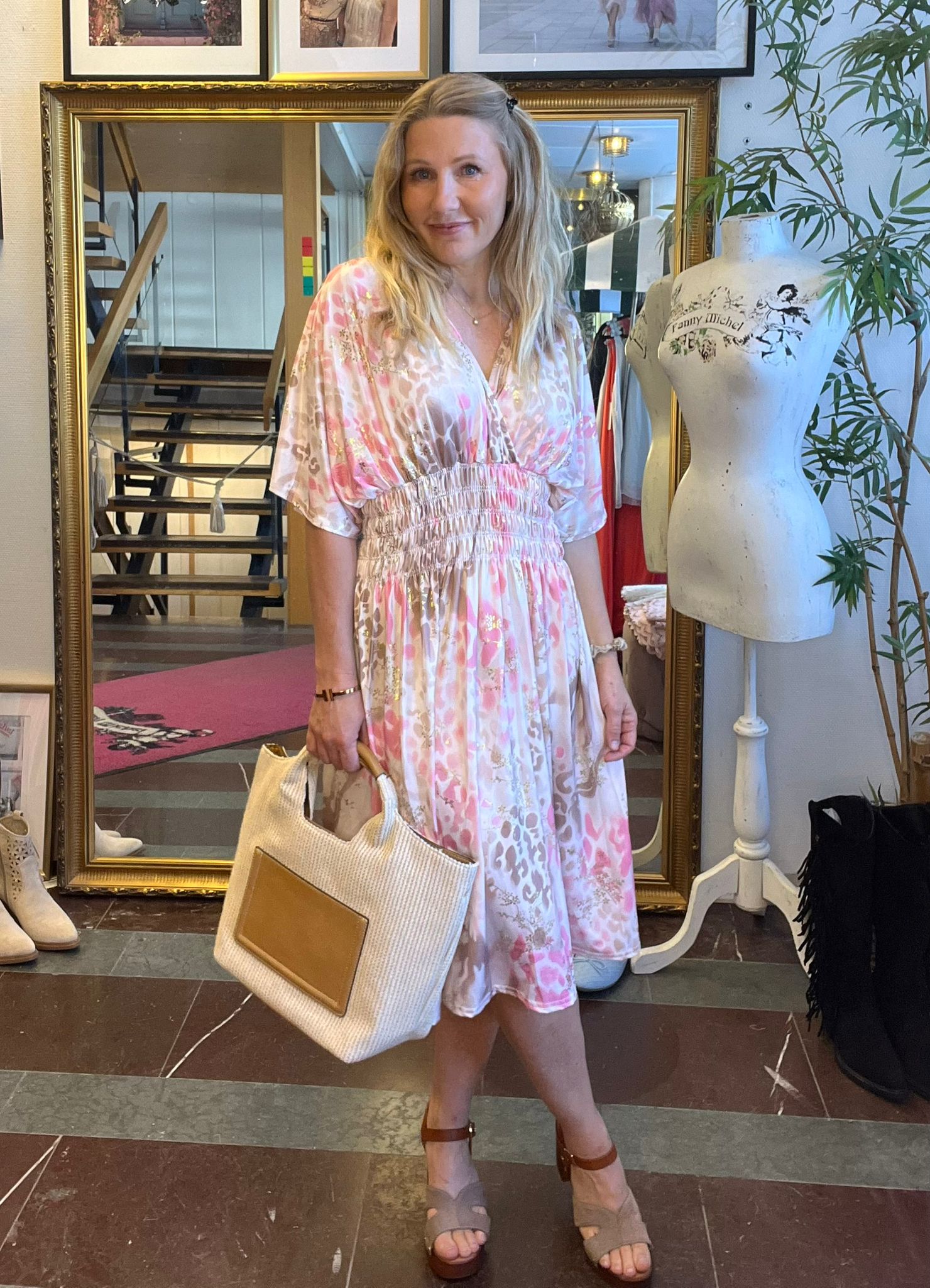 Leende kvinna poserar framför en stor guldramsspegel, klädd i rosa- och beigeblommig klänning och sandaler, och håller en stor beige handväska. Hon står i en butik med trappa, klädställning och provdocka med texten “Fanny Michel”.