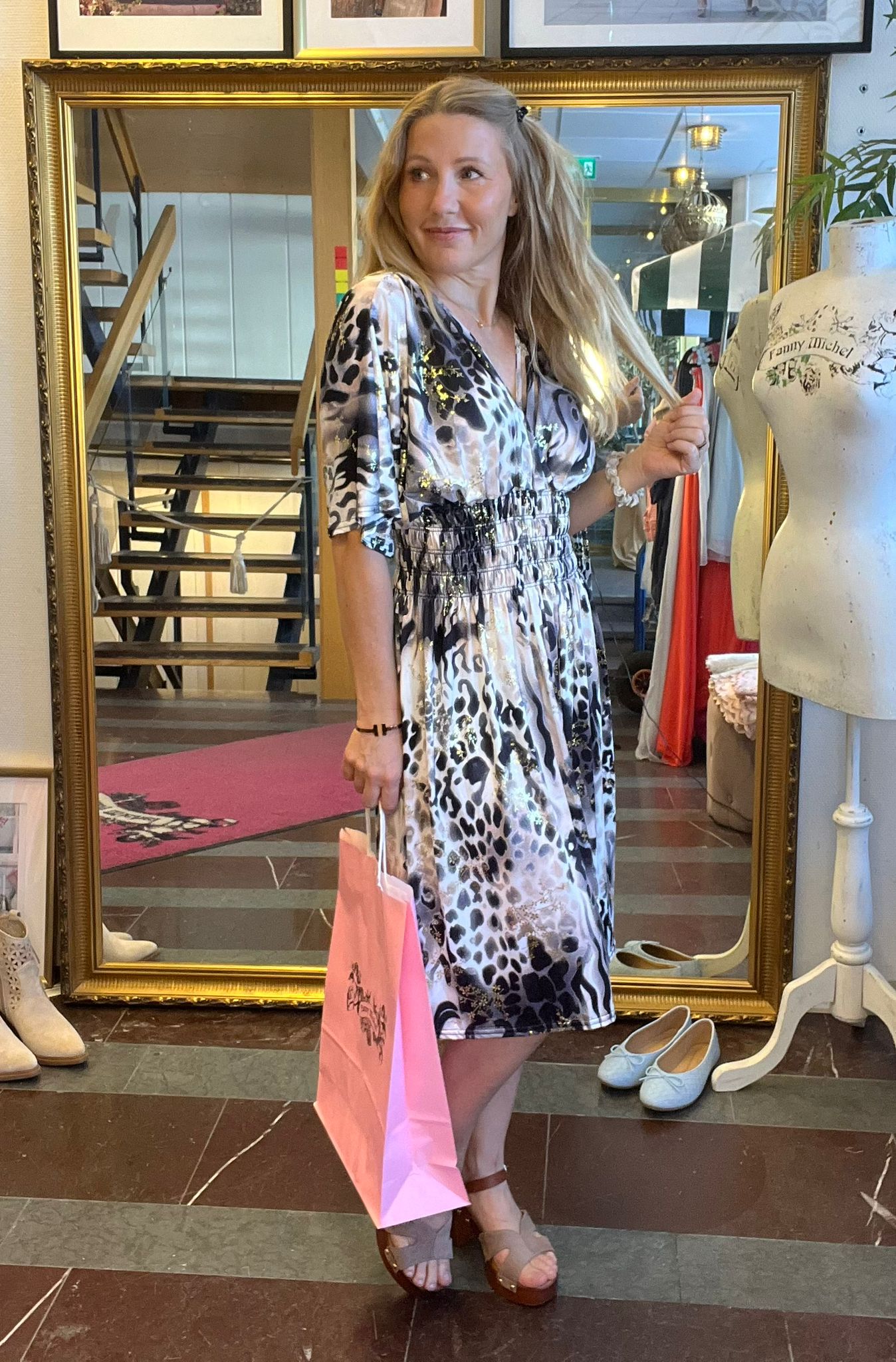 Kvinna i mönstrad klänning poserar framför en stor guldramsspegel, håller upp en hårslinga och bär en rosa shoppingpåse. Hon står i en klädbutik med provdocka märkt “Fanny Michel” och skor på golvet.