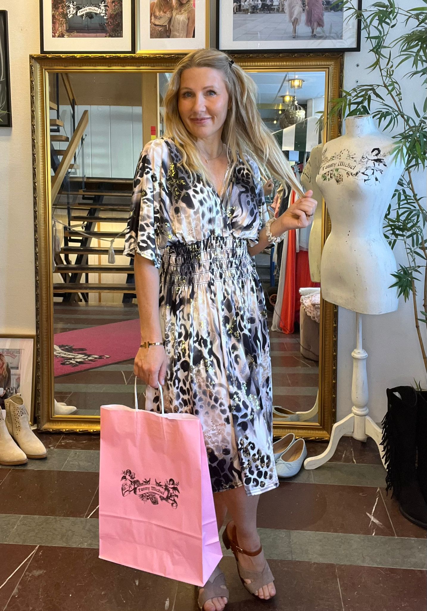 Leende kvinna i mönstrad klänning står i en butik och håller en rosa shoppingpåse med texten ”Fanny Michel”. Bakom henne syns en stor guldramsspegel, skor på golvet och en provdocka.