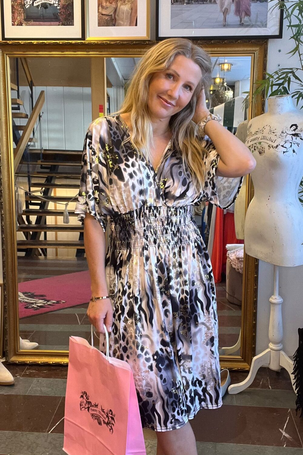 Leende kvinna i mönstrad klänning poserar med handen i håret och håller en rosa shoppingpåse. Hon står i en butik framför en stor guldramsspegel; bredvid syns en provdocka med texten ”Fanny Michel”.