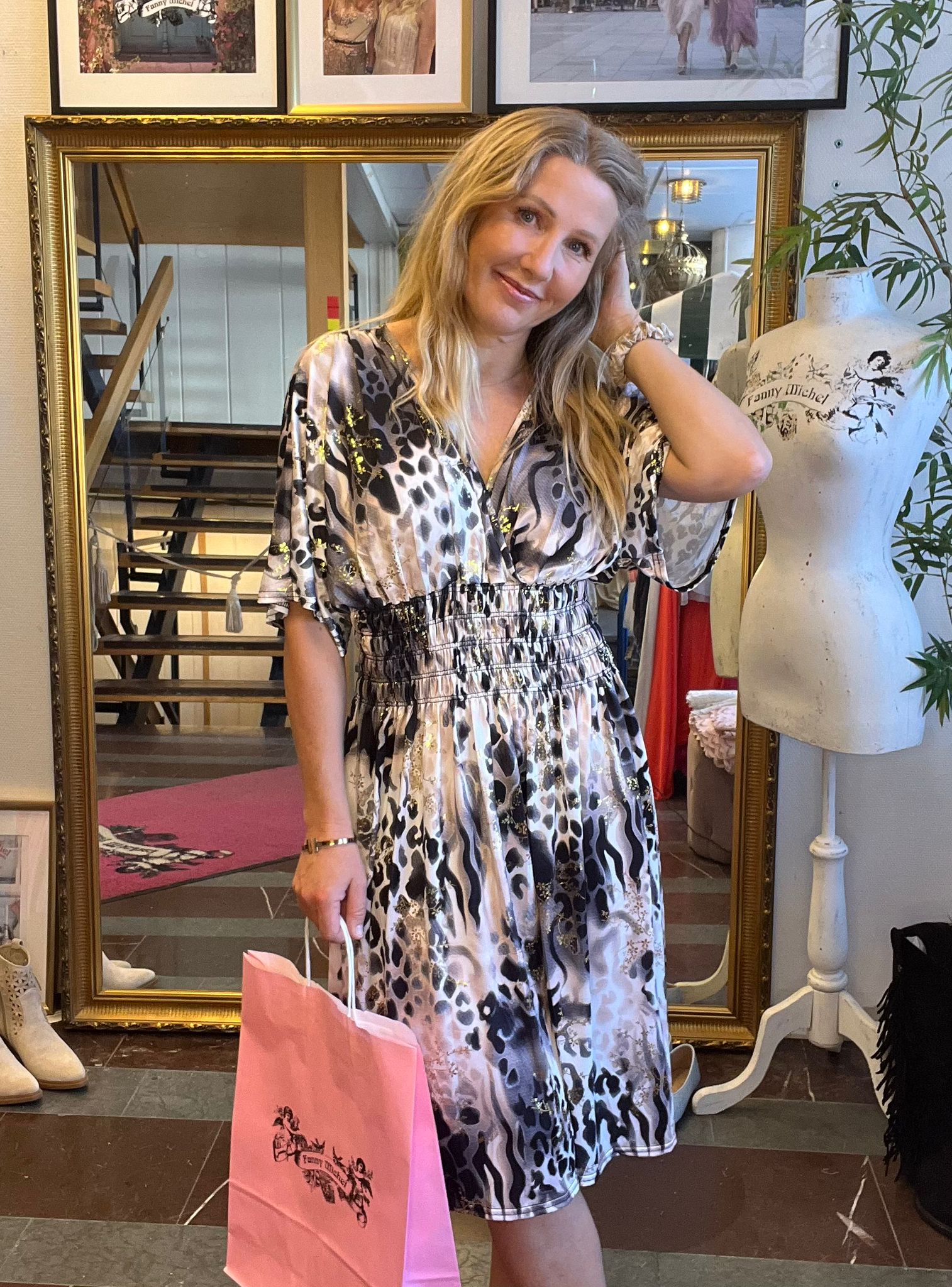 Leende kvinna i mönstrad klänning poserar med handen i håret och håller en rosa shoppingpåse. Hon står i en butik framför en stor guldramsspegel; bredvid syns en provdocka med texten ”Fanny Michel”.