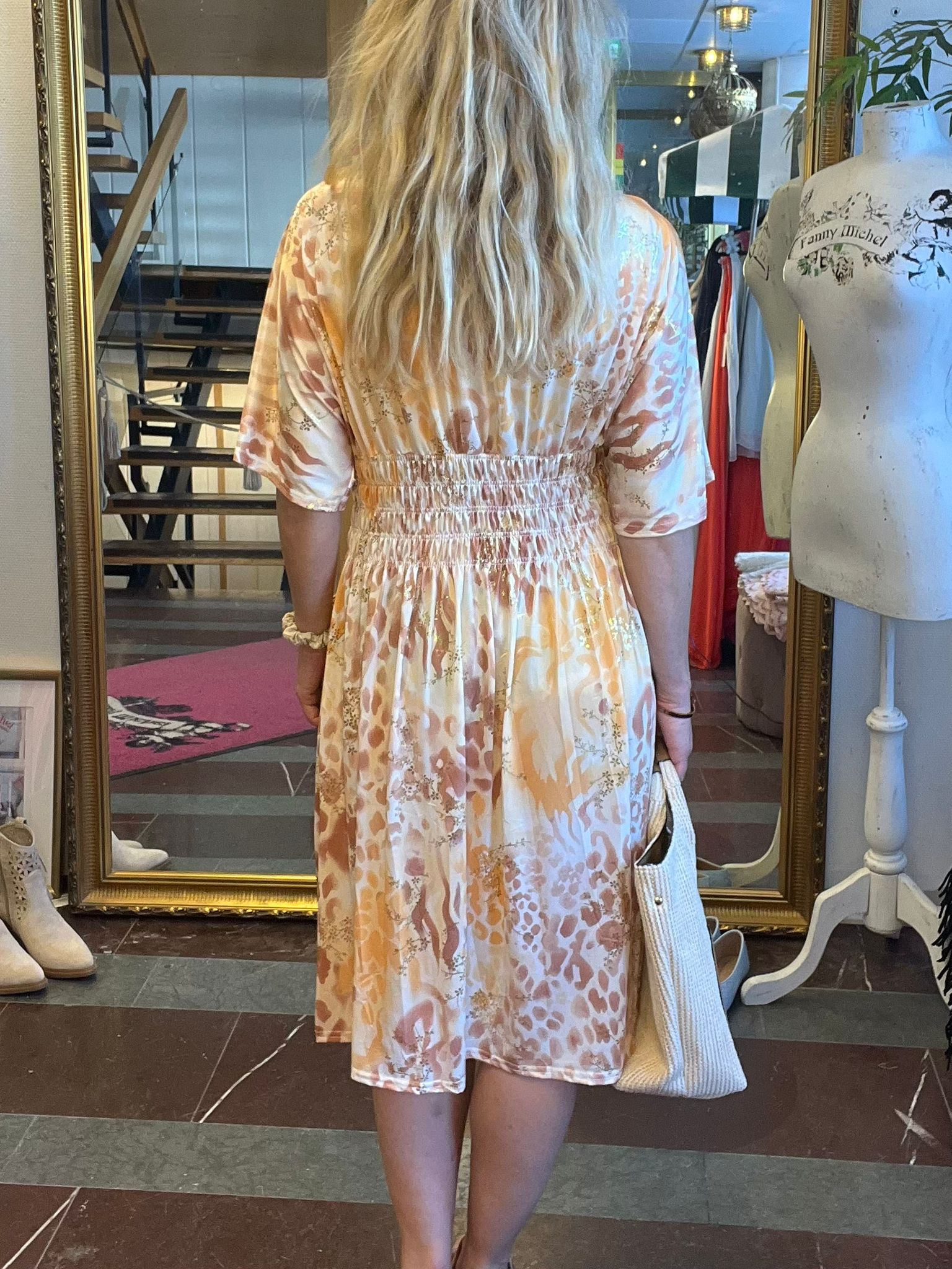 Person med långt blont hår står med ryggen mot kameran framför en stor guldramsspegel och provar en ljus, mönstrad klänning, med en vit stickad väska i handen, i en butik med klädställning och provdocka med texten “Fanny Michel”.