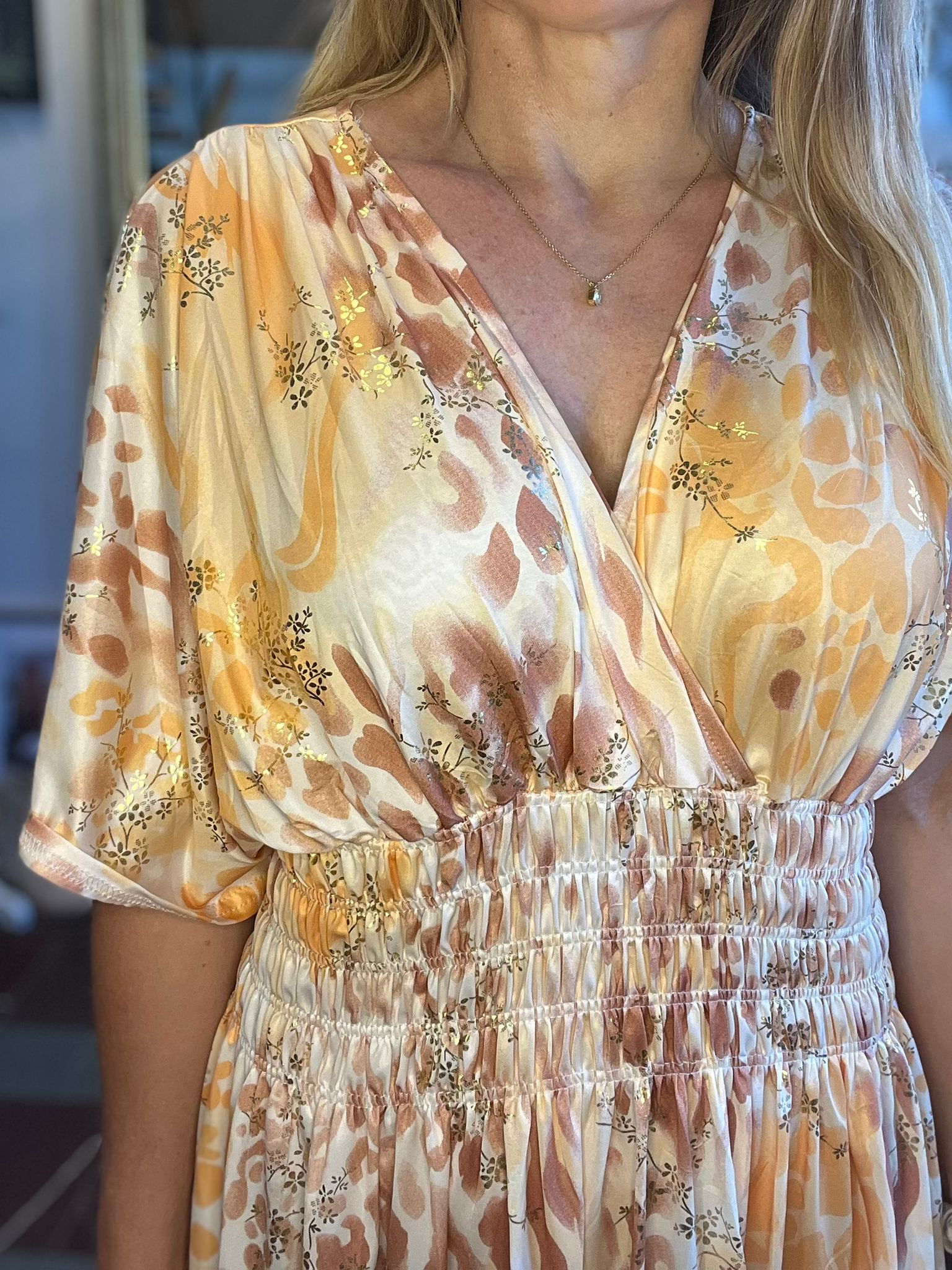 Kvinna syns från axlar till midja och bär en ljus omlottklänning med leopardlikt mönster och guldfärgade blomdetaljer. Hon står inomhus och har ett tunt halsband med liten droppformad sten.