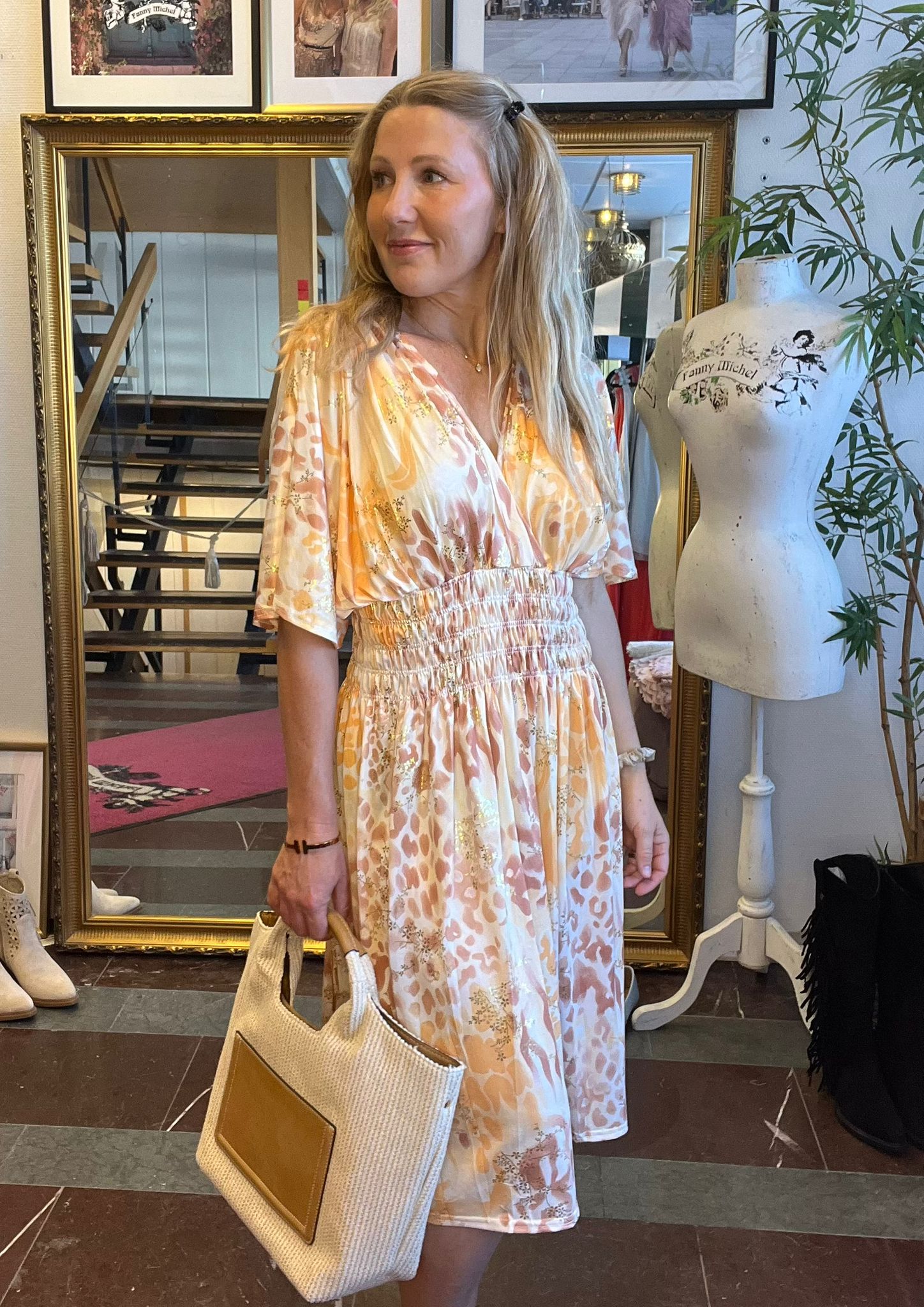 Kvinna i ljus, mönstrad klänning håller en stor stråväska och tittar åt sidan i en butik. Bakom henne står en guldramsspegel, trappa och provdocka med texten ”Fanny Michel”, samt en grön växt.