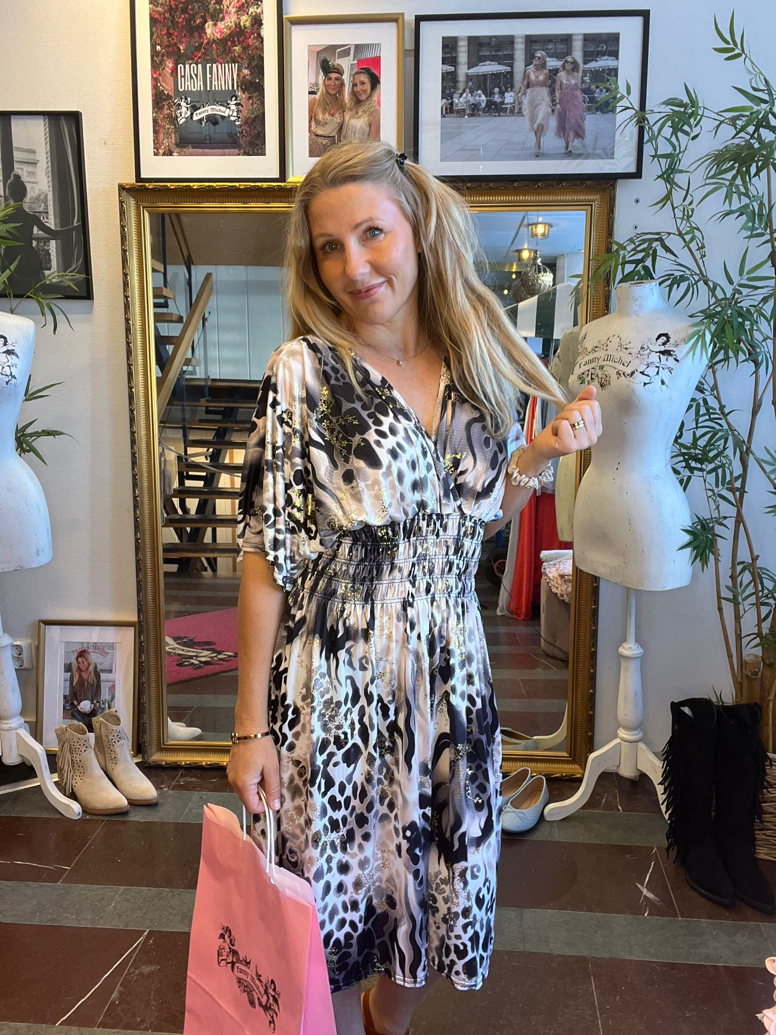 Kvinna i mönstrad klänning står och ler i en butik, håller en rosa shoppingpåse och drar i sitt hår. Bakom henne syns stor spegel, skyltdockor, skor och inramade foton med texten ”CASA FANNY”.