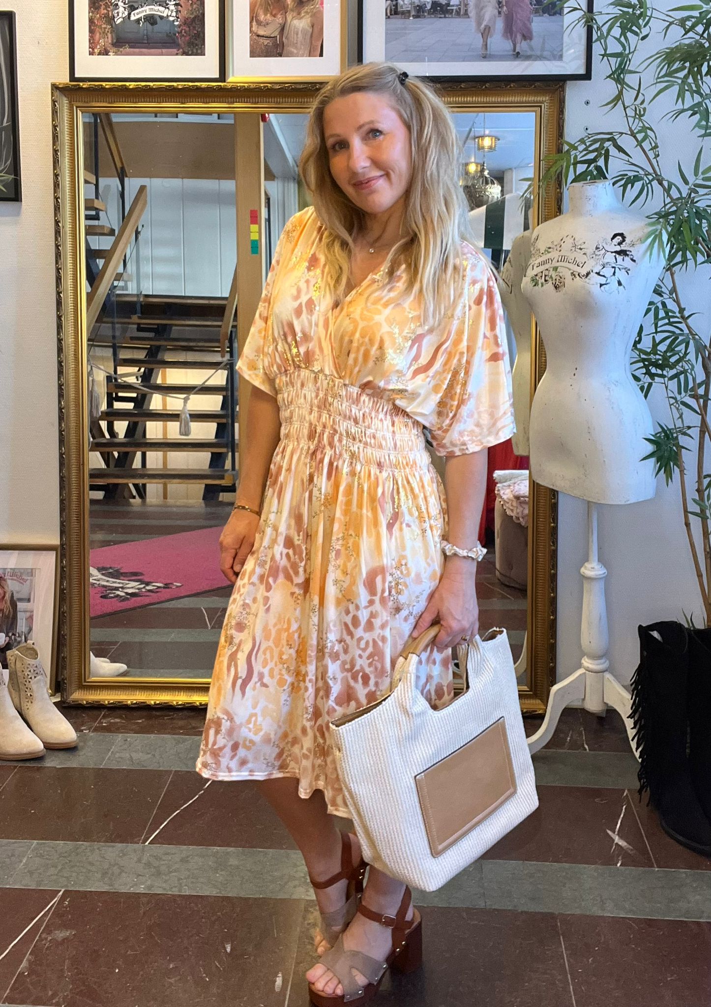 Leende kvinna står i en butiksmiljö och poserar i en ljus, mönstrad klänning medan hon håller en stor beige tygväska. Bakom henne syns en guldramsspegel, en trapp och en provdocka med texten ”Fanny Michel”.
