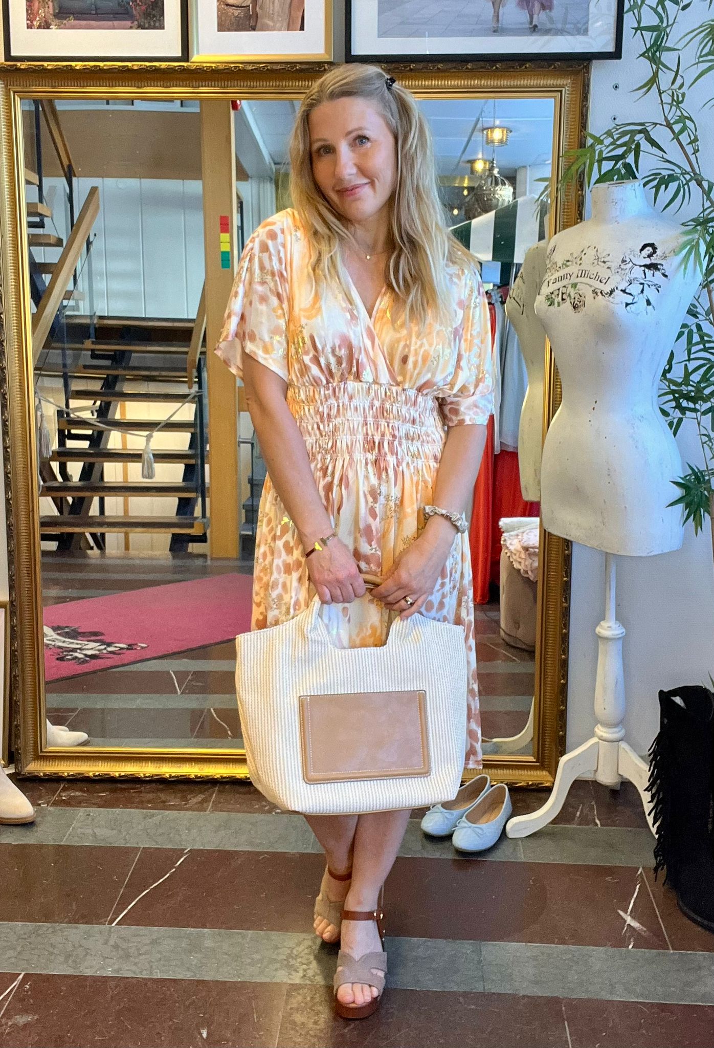 Blond kvinna i mönstrad ljus klänning står och ler medan hon håller en stor vit tygväska framför sig. Hon är i en butik med guldramsspegel, trappa och provdocka med texten “Fanny Michel”.