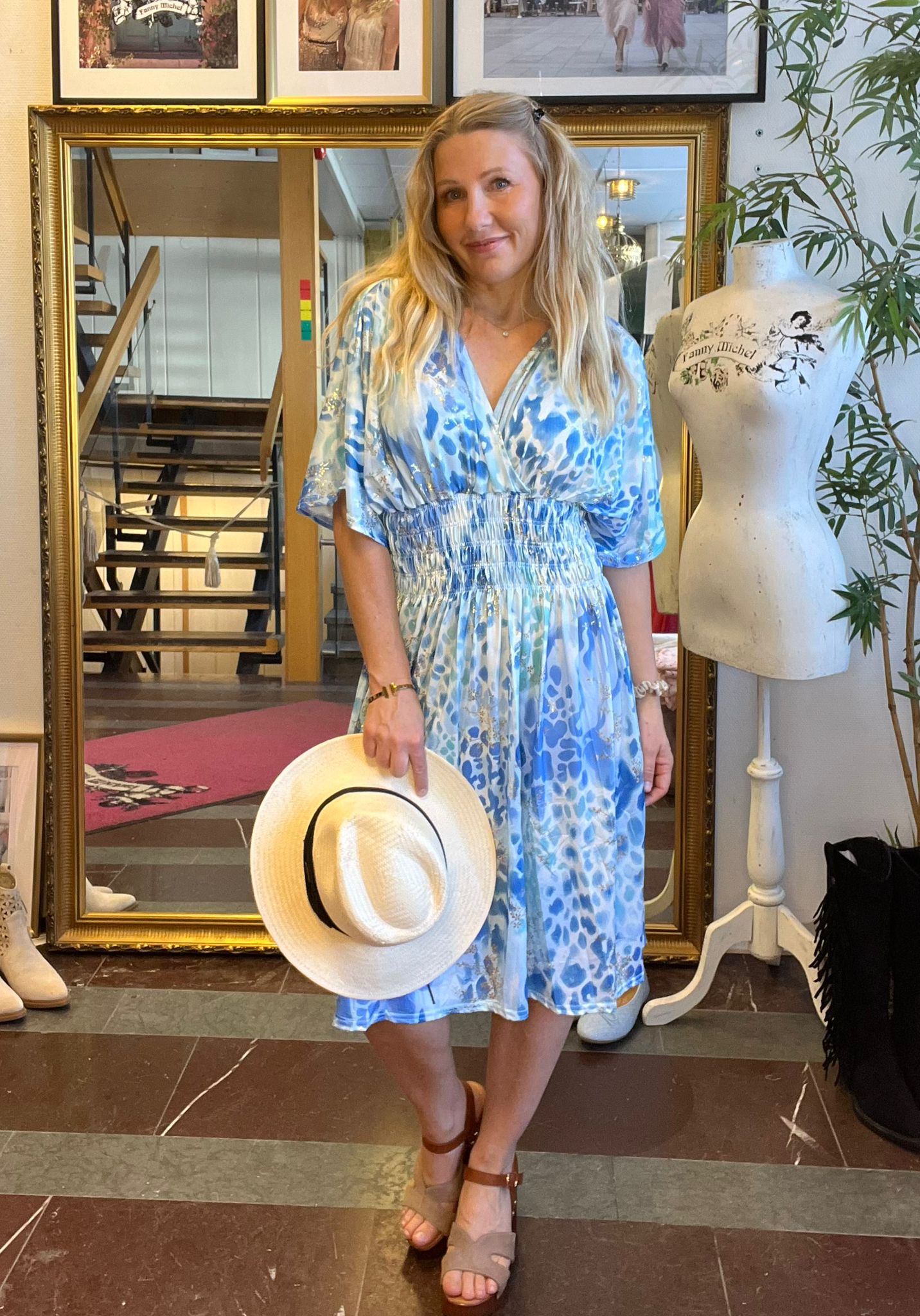 Blond kvinna i blåmönstrad klänning står och ler, håller en vit stråhatt med svart band. Hon står framför en stor guldramsspegel i en butik, bredvid provdocka med texten “Fanny Michel”.