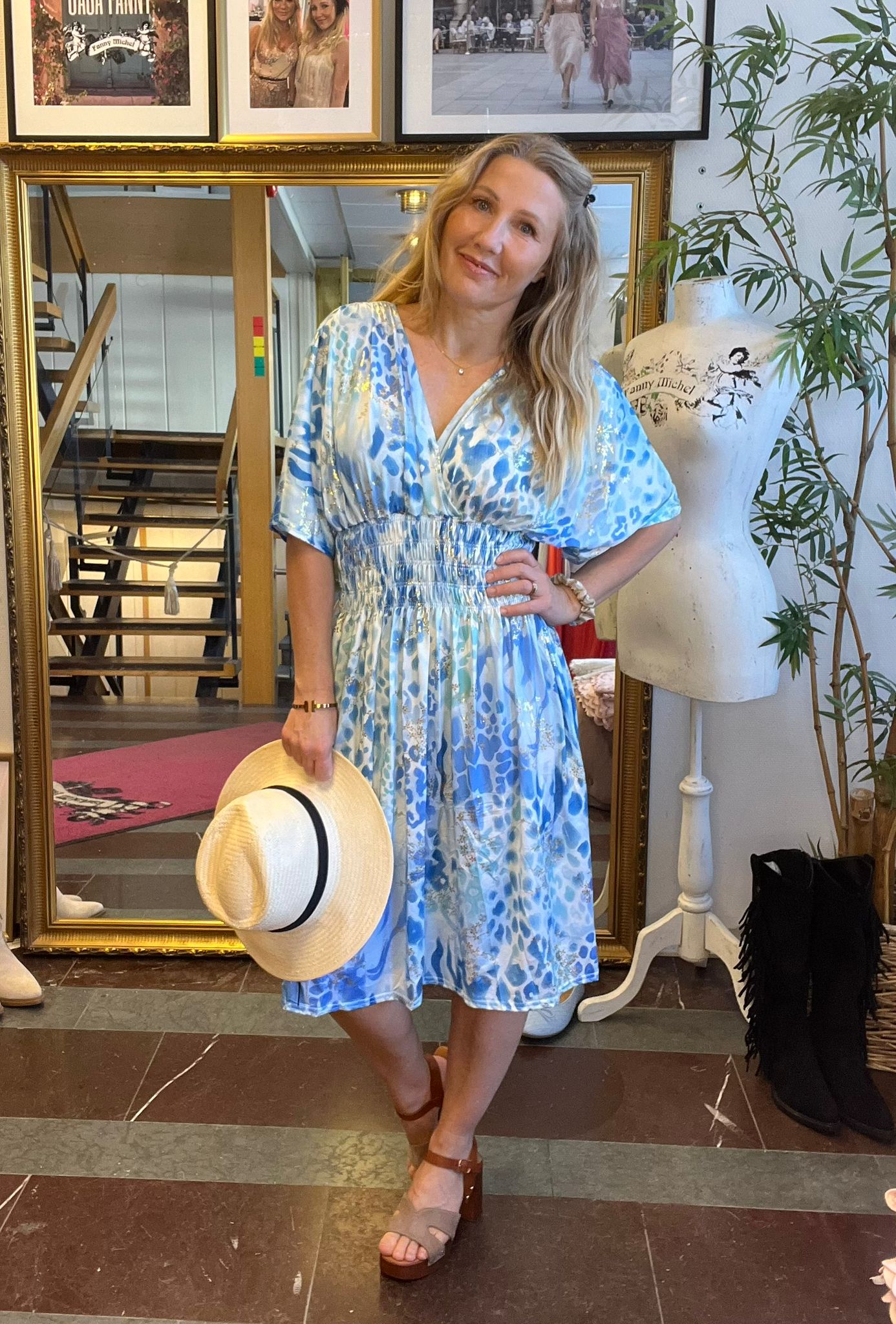 Kvinna poserar med handen på höften och håller en halmhatt, klädd i blåvit mönstrad klänning. Hon står framför en stor guldramsspegel i en butik, intill en provdocka med texten “Fanny Michel”.