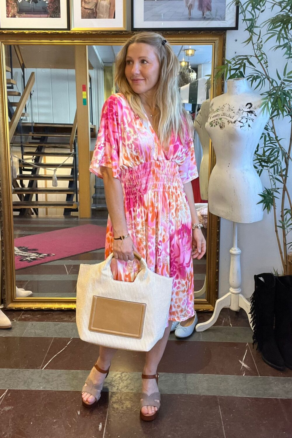 Kvinna i rosa mönstrad klänning står och ler i en klädbutik, håller en stor vit strandväska och tittar åt sidan. Bakom henne syns guldramsspegel, trappa och provdocka med texten “Fanny Michel”.