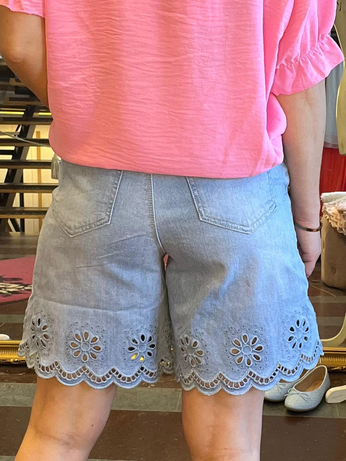 Person sedd bakifrån står inomhus med en rosa topp och ljusblå denimshorts med broderad spetskant och blommönster. I bakgrunden syns en spegel, skor på golvet och möbler.