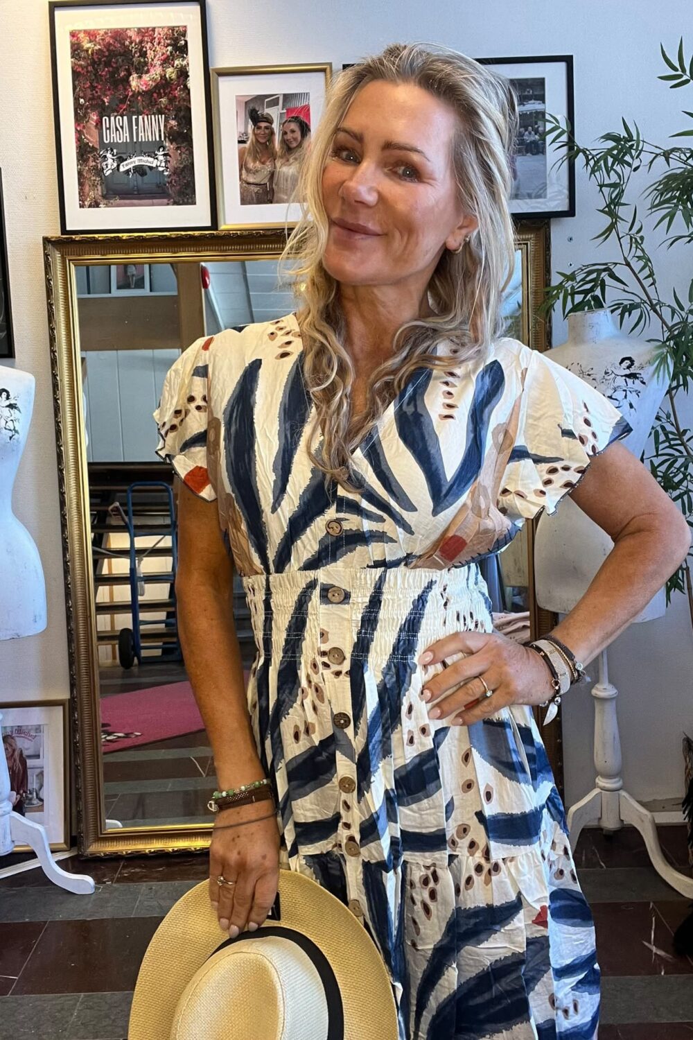 Leende kvinna poserar med handen på höften och håller en stråhatt i en butik eller ateljé. Hon bär en mönstrad klänning. Bakom syns spegel, skyltdockor och inramade bilder med texten ”CASA FANNY”.