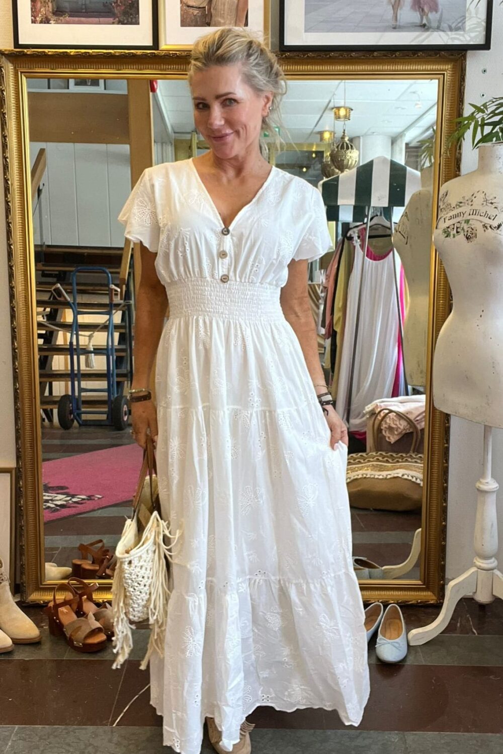 Kvinna står och poserar framför en stor guldramsspegel i en butik, iklädd vit broderad maxiklänning och hållande en fransad väska. Runt henne syns skor, klädställning och provdocka; på väggen står texten “CASA FANNY”.