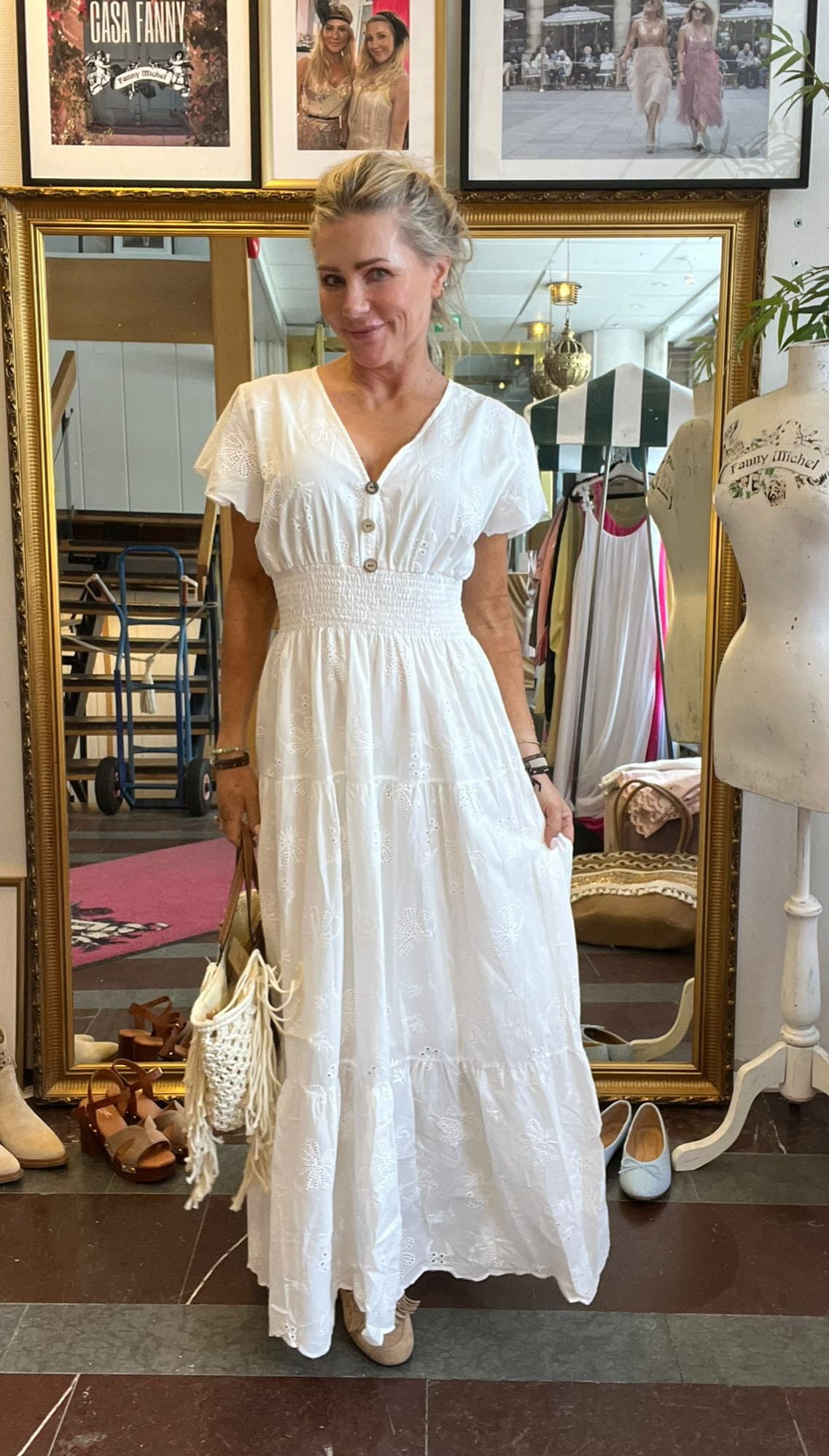 Kvinna står och poserar framför en stor guldramsspegel i en butik, iklädd vit broderad maxiklänning och hållande en fransad väska. Runt henne syns skor, klädställning och provdocka; på väggen står texten “CASA FANNY”.