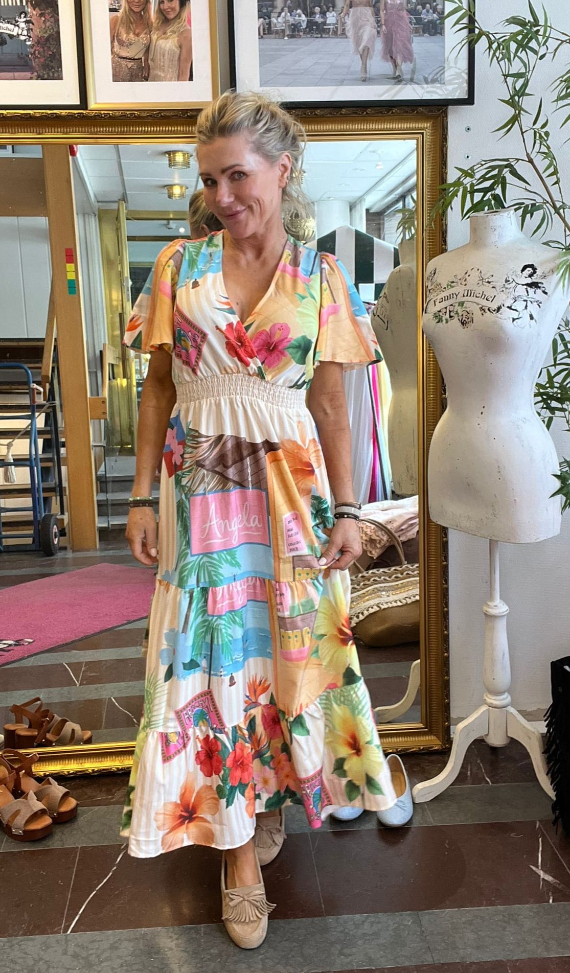 Leende kvinna står framför en stor guldramsspegel och visar upp en färgstark, blommig långklänning. I rummet syns provdocka med texten “Fanny Michel”, inramade foton på väggen samt skor på golvet.