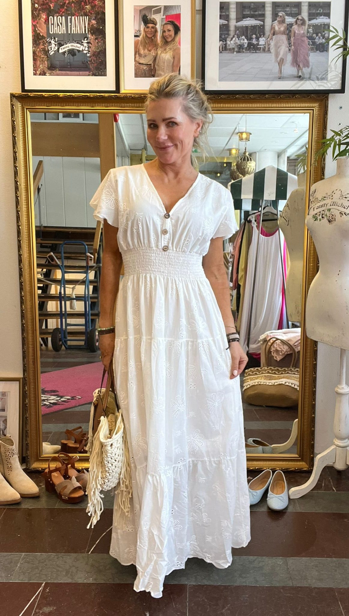 Kvinna i vit broderad maxiklänning står och ler framför en stor guldramsspegel i en klädbutik, håller en fransig handväska. Skor och klädhängare syns runtom. På väggbilderna står texten “CASA FANNY” och “Fanny Michel”.