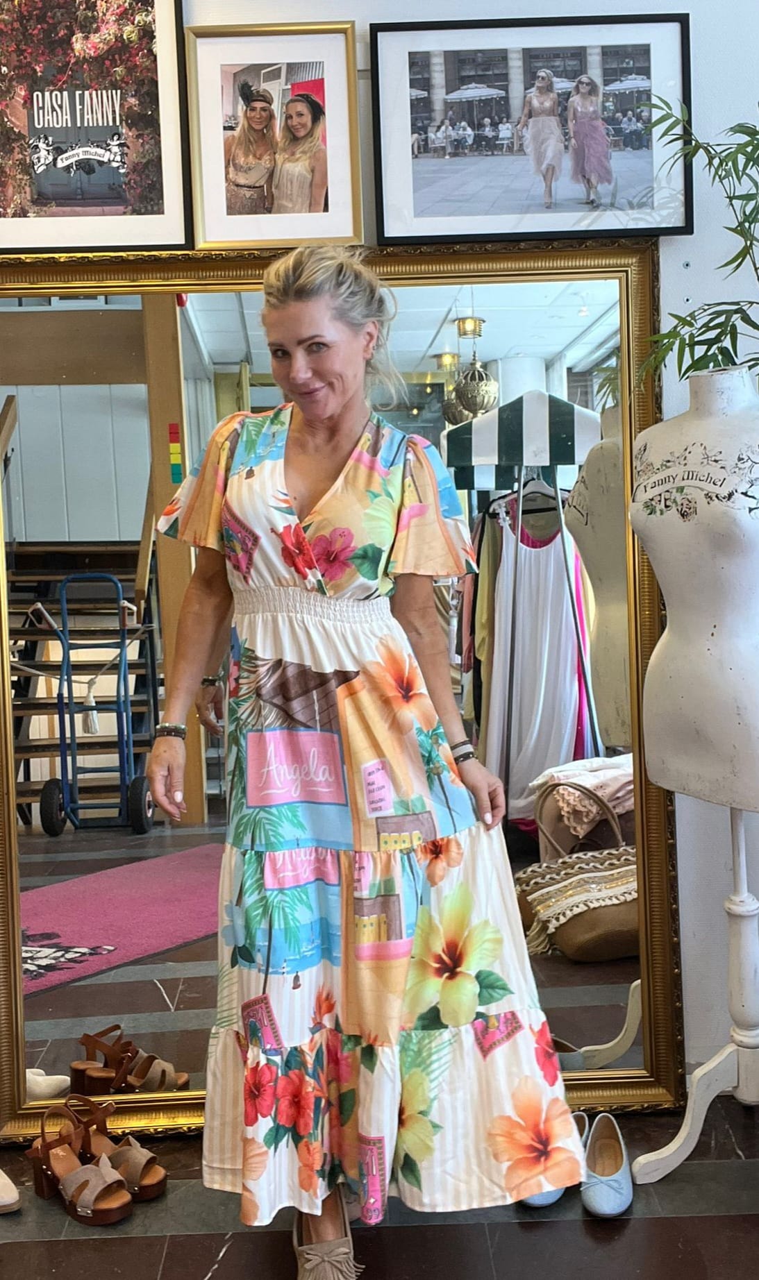 Leende kvinna poserar i en butik/ateljé framför en stor spegel, iklädd en färgstark blommig klänning med texten “Angela”. Runt henne syns skor, klädställning och en provdocka; på väggen hänger inramade foton och skylten “CASH FANNY”.
