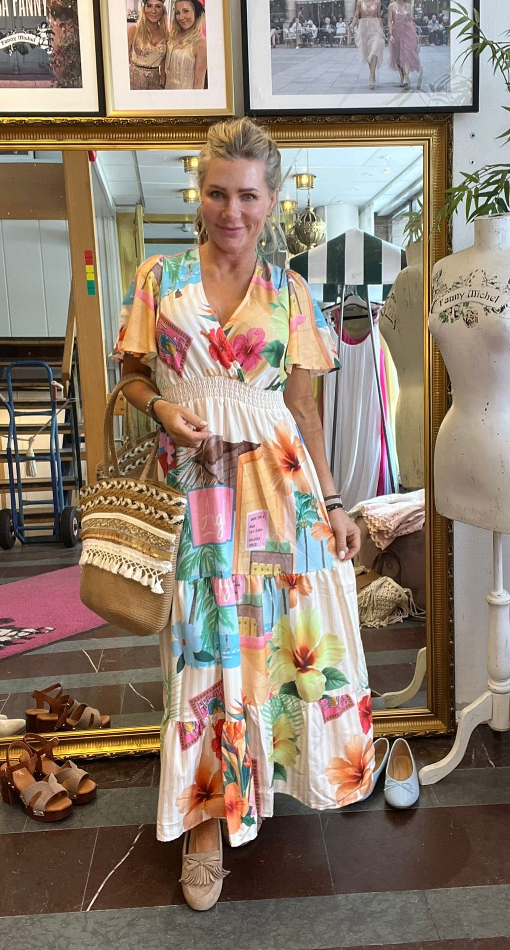 Kvinna i färgglad blommig maxiklänning står och poserar framför en stor guldramad spegel i en butik, med stråväska över axeln. I bakgrunden syns kläder, skor och en provdocka med texten “Fanny Michel”.