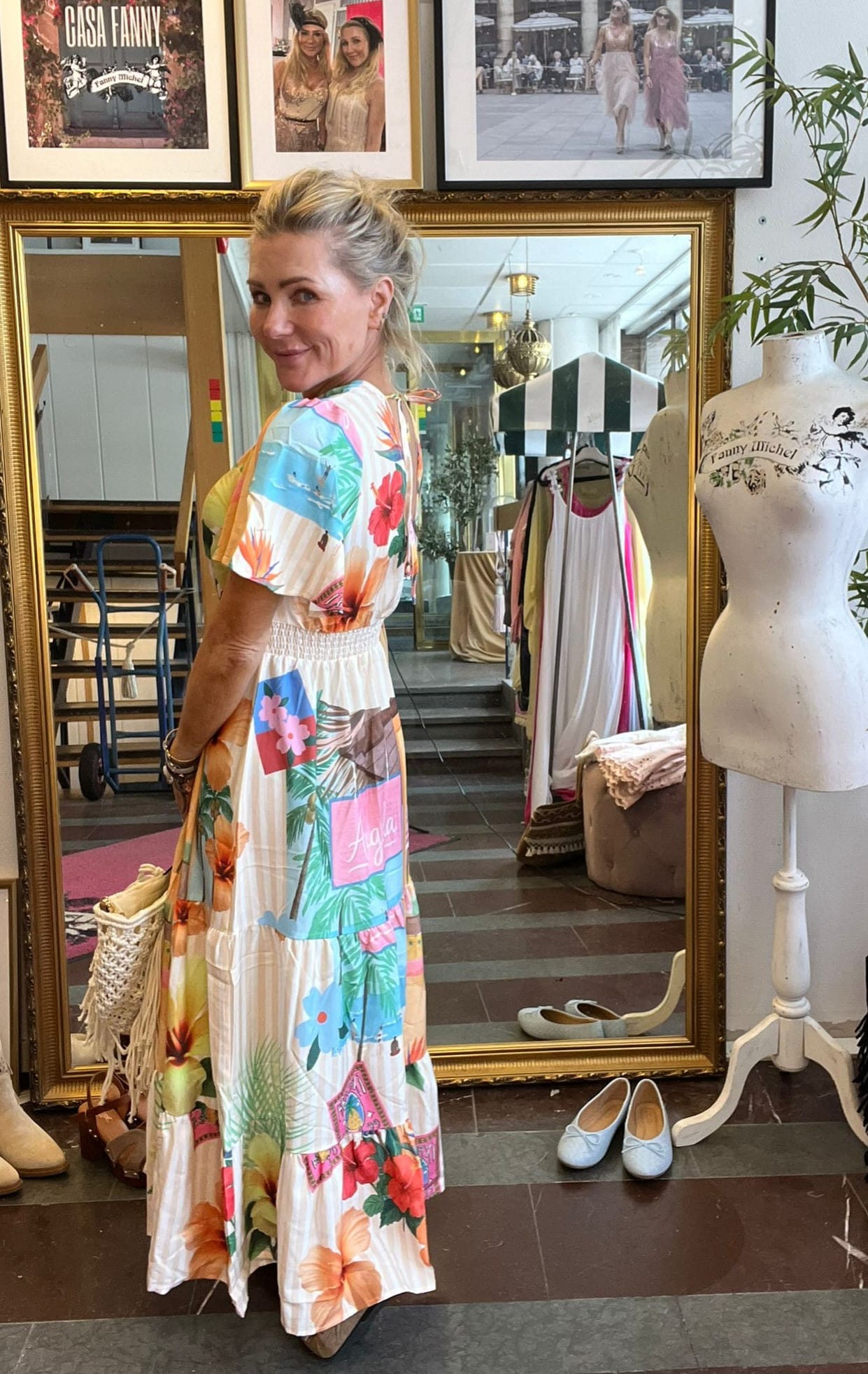 Blond kvinna står framför en stor guldramad spegel och ler över axeln, iklädd en färgstark mönstrad maxiklänning. Runt henne syns klädställning och skor; en provdocka med texten “Fanny Michel” och väggbilder ovanför.