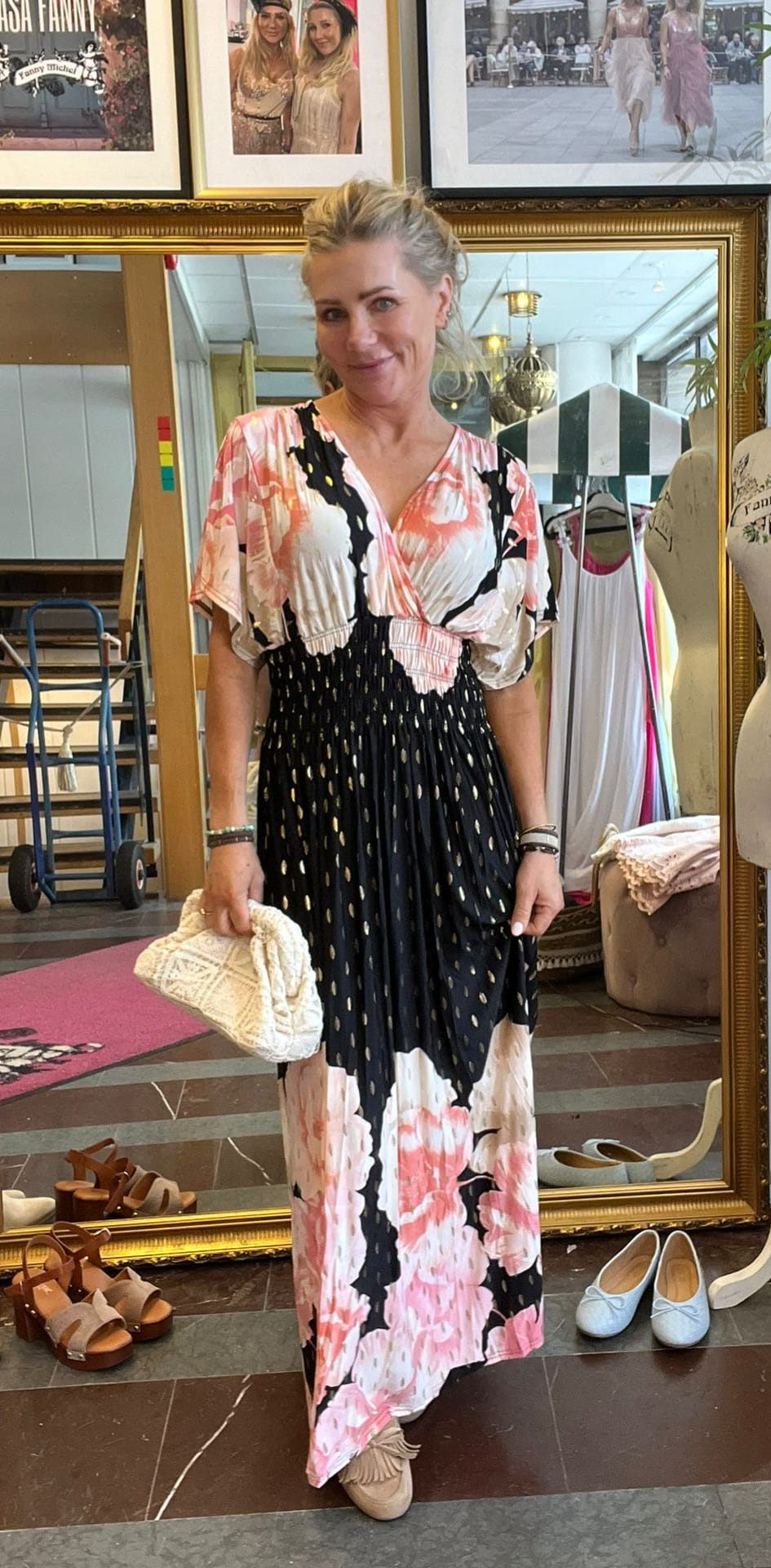 Blond kvinna poserar framför en stor guldramsspegel och håller en vit virkad clutch. Hon bär en lång svart‑rosa blommig klänning. I bakgrunden syns klädställning, provdocka, skor på golvet och inramade foton.