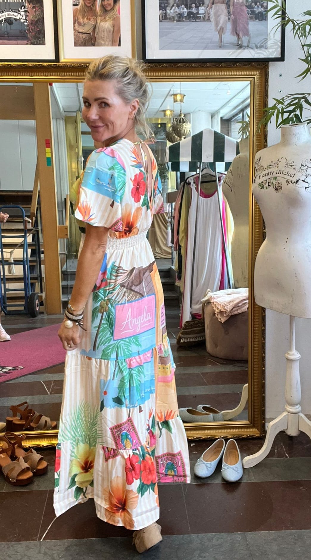Blond kvinna står vänd åt sidan och ler mot kameran, iklädd lång, färgstark klänning med blommor och texten “Angela”. Hon är i en butik/ateljé med stor guldramsspegel, skyltdocka och skor på golvet.