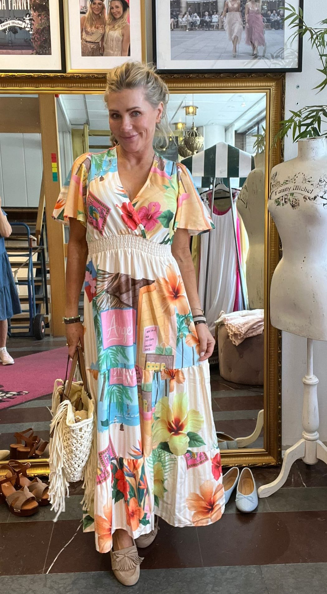 Kvinna står och poserar leende i en butik/ateljé, iförd en färgstark blommig långklänning och med en fransad handväska. Hon står framför en stor spegel, omgiven av skor, klädställning och provdocka med texten ”Fanny Michel”.