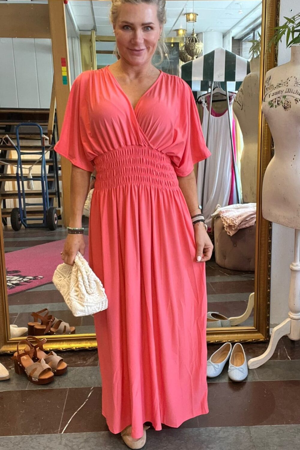 Blond kvinna står och poserar framför en stor guldramsspegel i en butik, klädd i en korallrosa, lång klänning och håller en vit virkad handväska. Skor och en provdocka syns runt henne.
