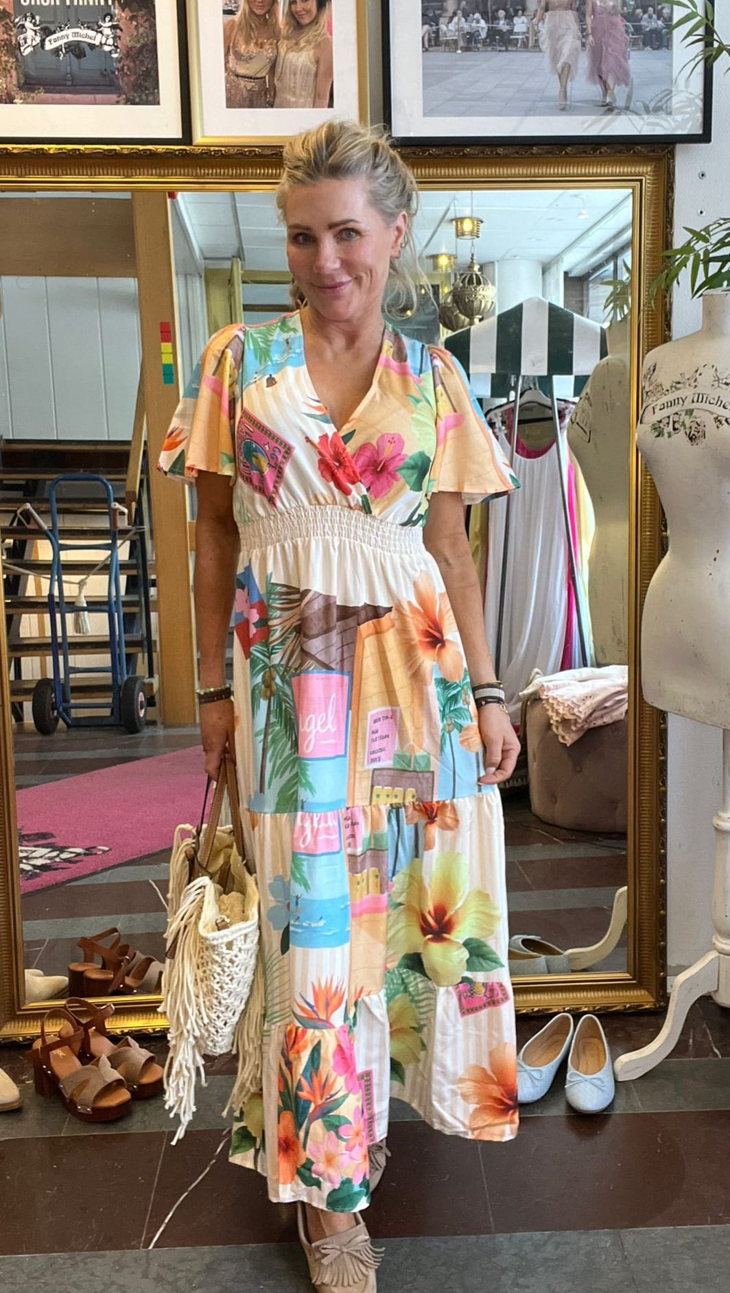 Kvinna står och poserar framför en stor guldramsspegel i en butik, iklädd lång, färgstark blommig klänning och håller en fransad, virkad väska. Runt henne syns skor på golvet och en provdocka.