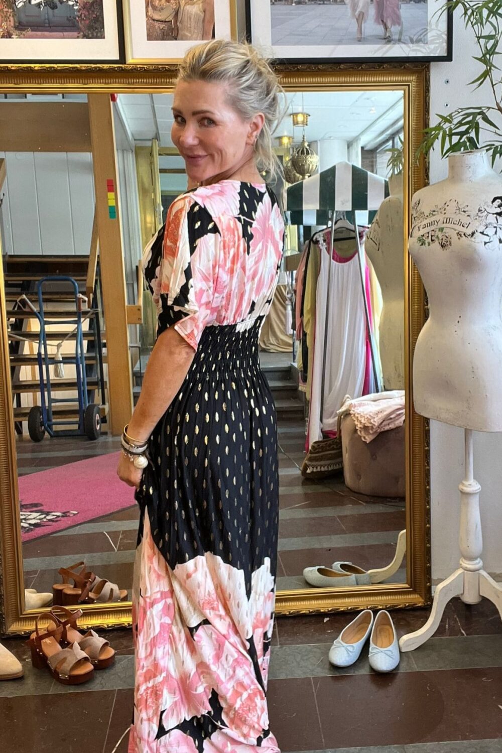 Blond kvinna vänder sig om och ler mot kameran medan hon provar en lång klänning med rosa blommönster och svart kjoldel med gulddetaljer, framför en stor spegel i en klädbutik med skor och provdocka. Text: “Fanny Michel”.