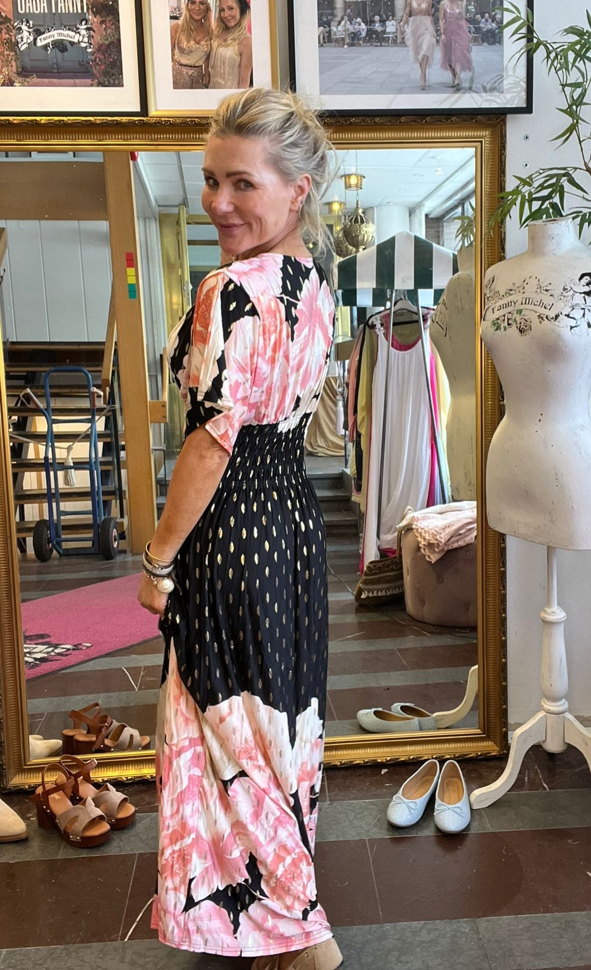 Blond kvinna vänder sig om och ler mot kameran medan hon provar en lång klänning med rosa blommönster och svart kjoldel med gulddetaljer, framför en stor spegel i en klädbutik med skor och provdocka. Text: “Fanny Michel”.