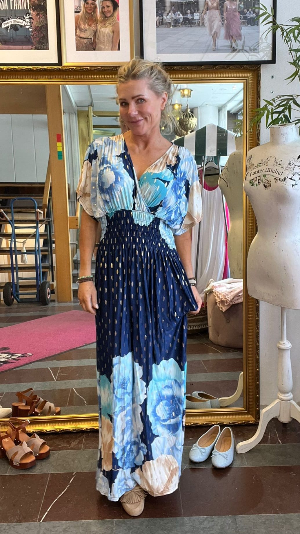 Blond kvinna står och ler framför en stor guldramsspegel och visar en lång blå klänning med stora blommor. Runt henne syns en provdocka med texten “Fanny Michel”, skor på golvet och inramade foton ovanför.