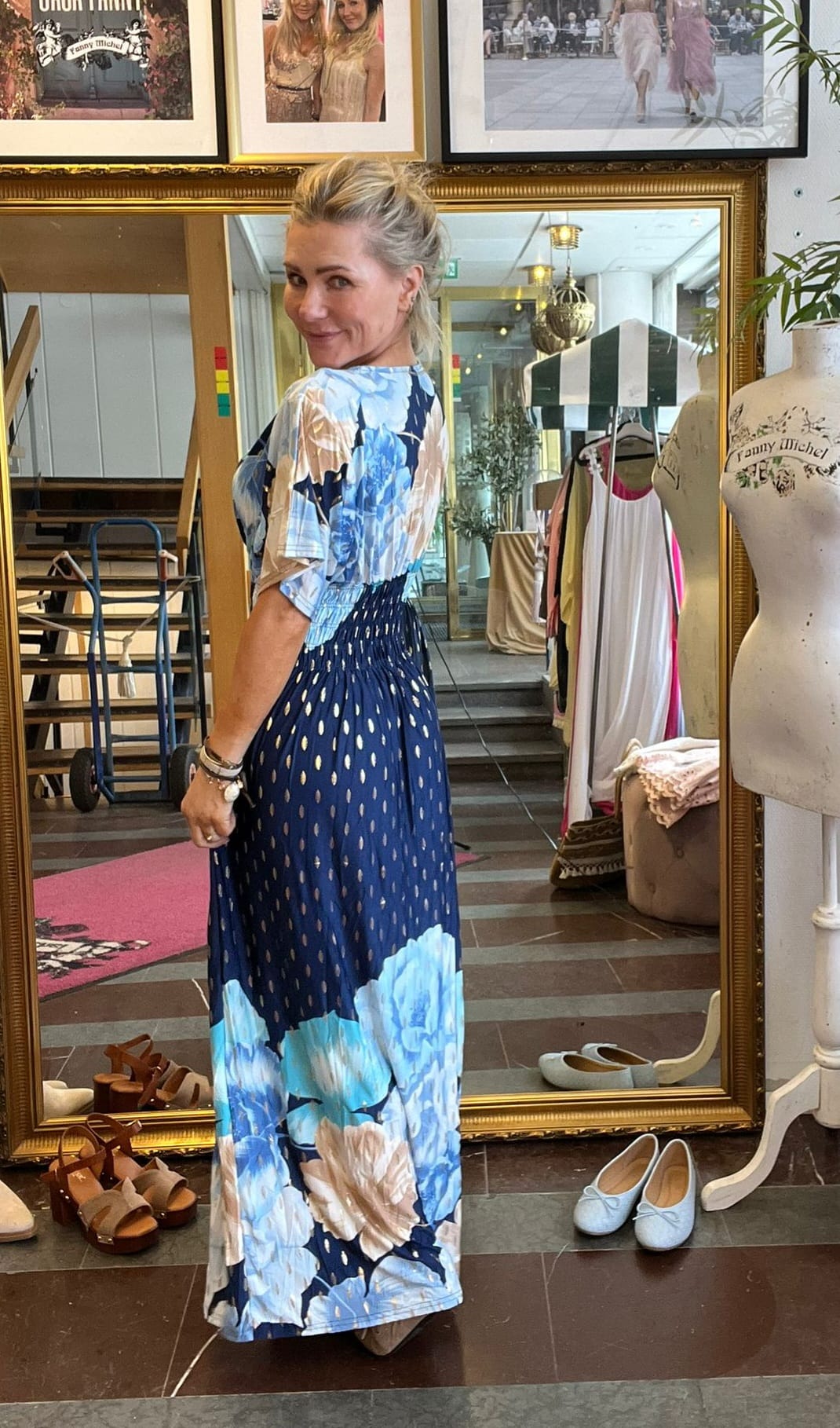 Blond kvinna poserar och tittar över axeln framför en stor guldramsspegel, iklädd lång blå klänning med stora ljusblå blommor. I bakgrunden syns klädhängare, skor på golvet och provdocka med texten “TILDE”.