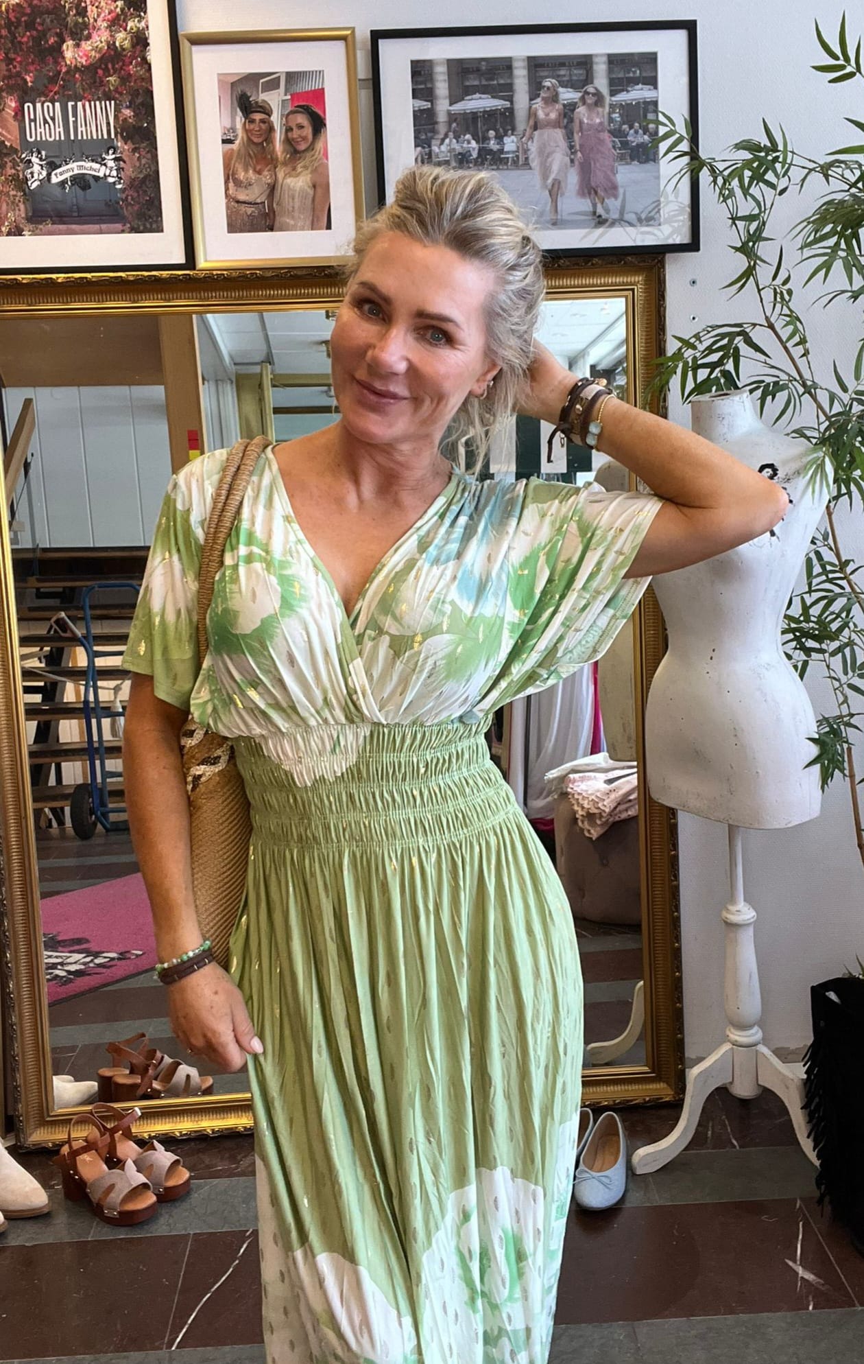 Kvinna i grönvit, mönstrad långklänning poserar och ler, med ena handen i håret och en stråväska över axeln, i en butik/ateljé med skor på golvet, skyltdocka och inramade foton; text: “CASA FANNY”.