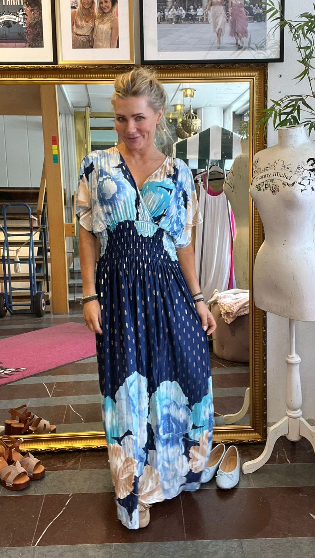Blond kvinna står och ler i en butik/ateljé och håller lätt i sin långa blå klänning med stora ljusblå blommor. Runt henne syns en stor guldramsspegel, skor på golvet och provdocka med texten “Fanny Michel”.