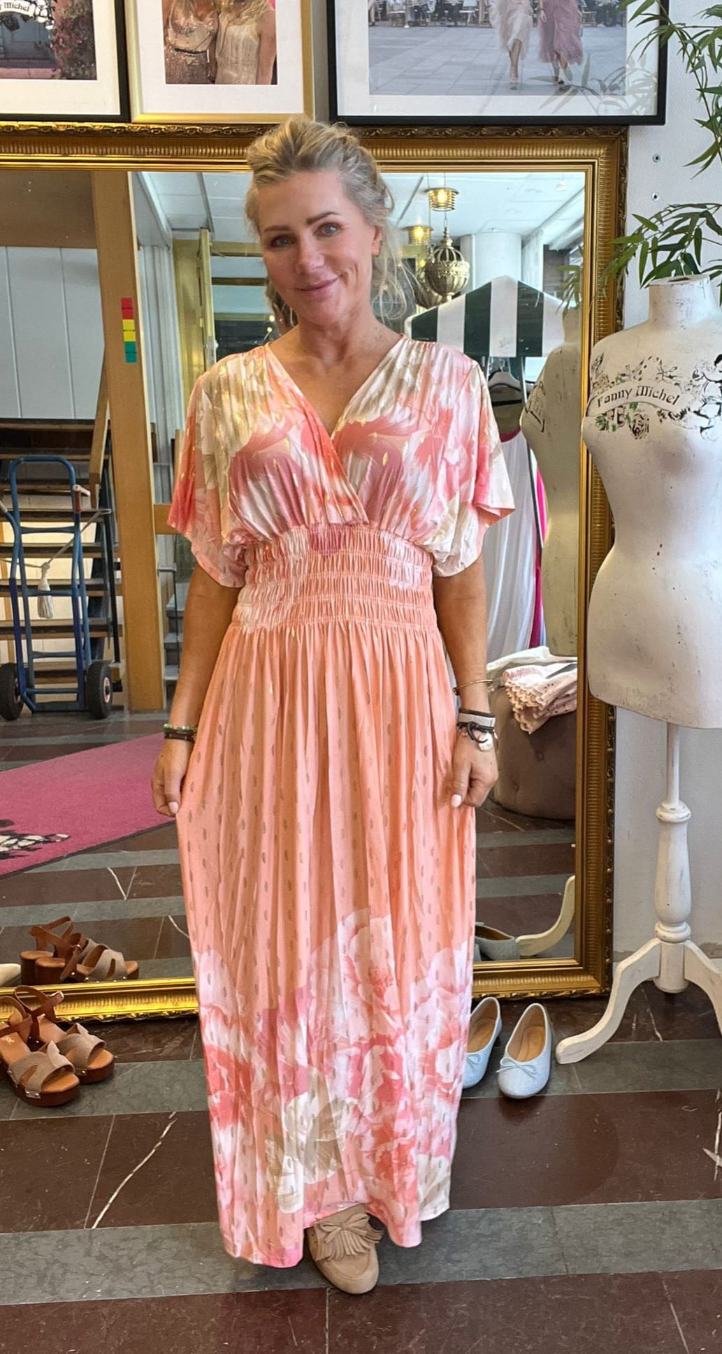 Kvinna står och ler framför en stor guldramsspegel och visar upp en lång rosa och vit mönstrad klänning. Hon är i en butik med skor på golvet och en provdocka med texten “Fanny Michel”.