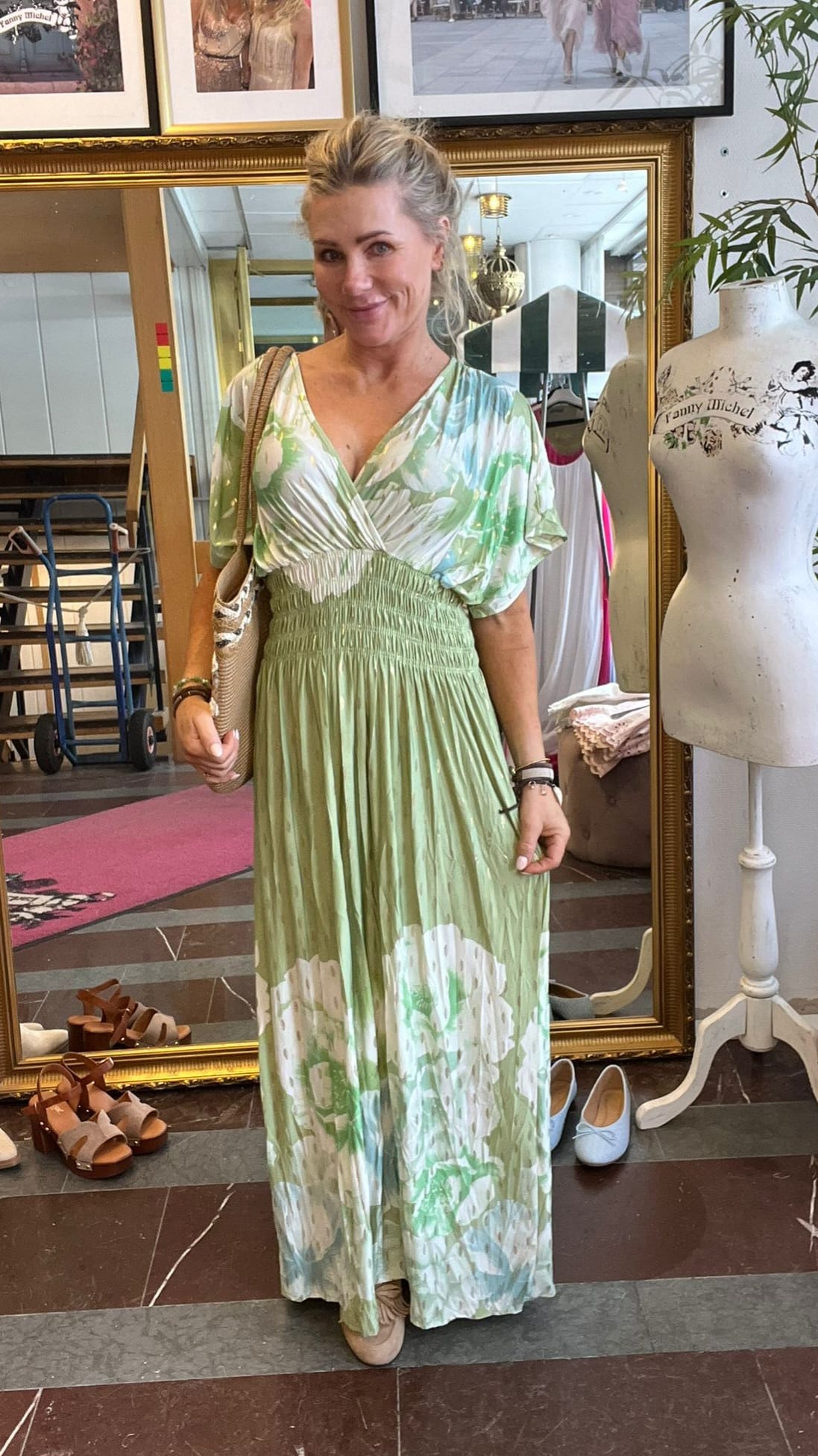 Leende kvinna står framför en stor guldramsspegel och poserar i en lång grönvit mönstrad klänning med axelväska. Runt henne syns skor på golvet, en provdocka med texten ”Fanny Michel” och butiksmiljö.