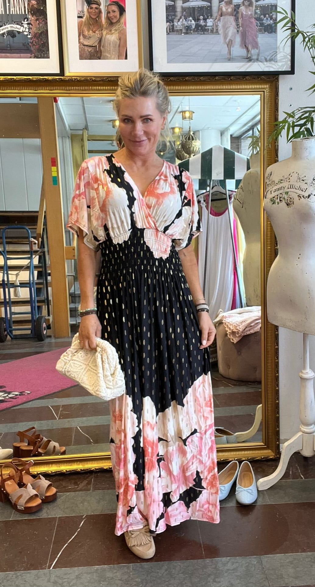 Blond kvinna poserar och håller en vit flätad clutch, iklädd lång mönstrad klänning i svart, rosa och vitt. Hon står framför en stor guldramsspegel i en butik, med skor på golvet och en provdocka märkt “Fanny Michel”.